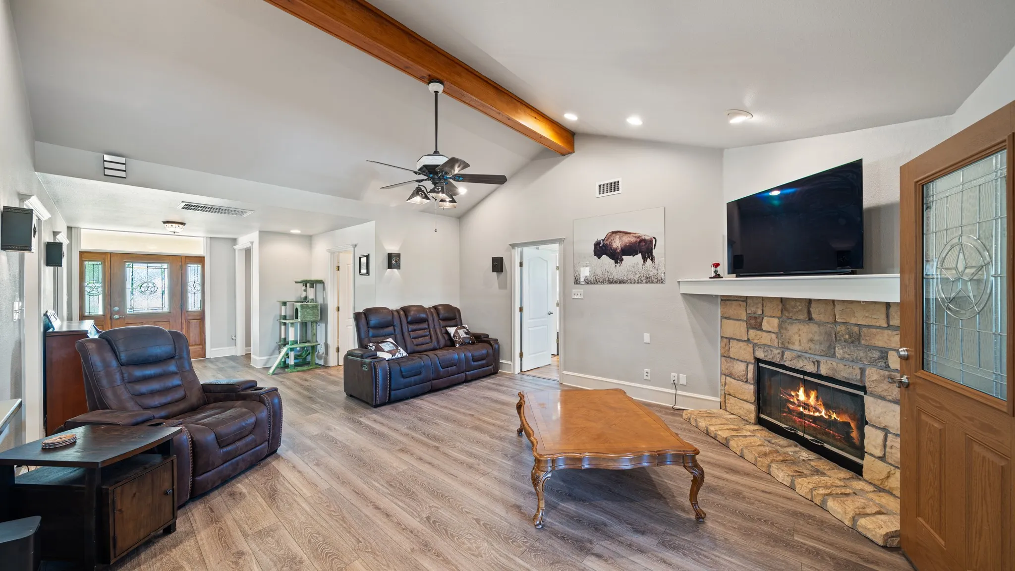 Vaulted ceiling with wood beam, wood-finish flooring, stone-faced fireplace with hearth, ceiling fan with light fixture, and recessed lighting