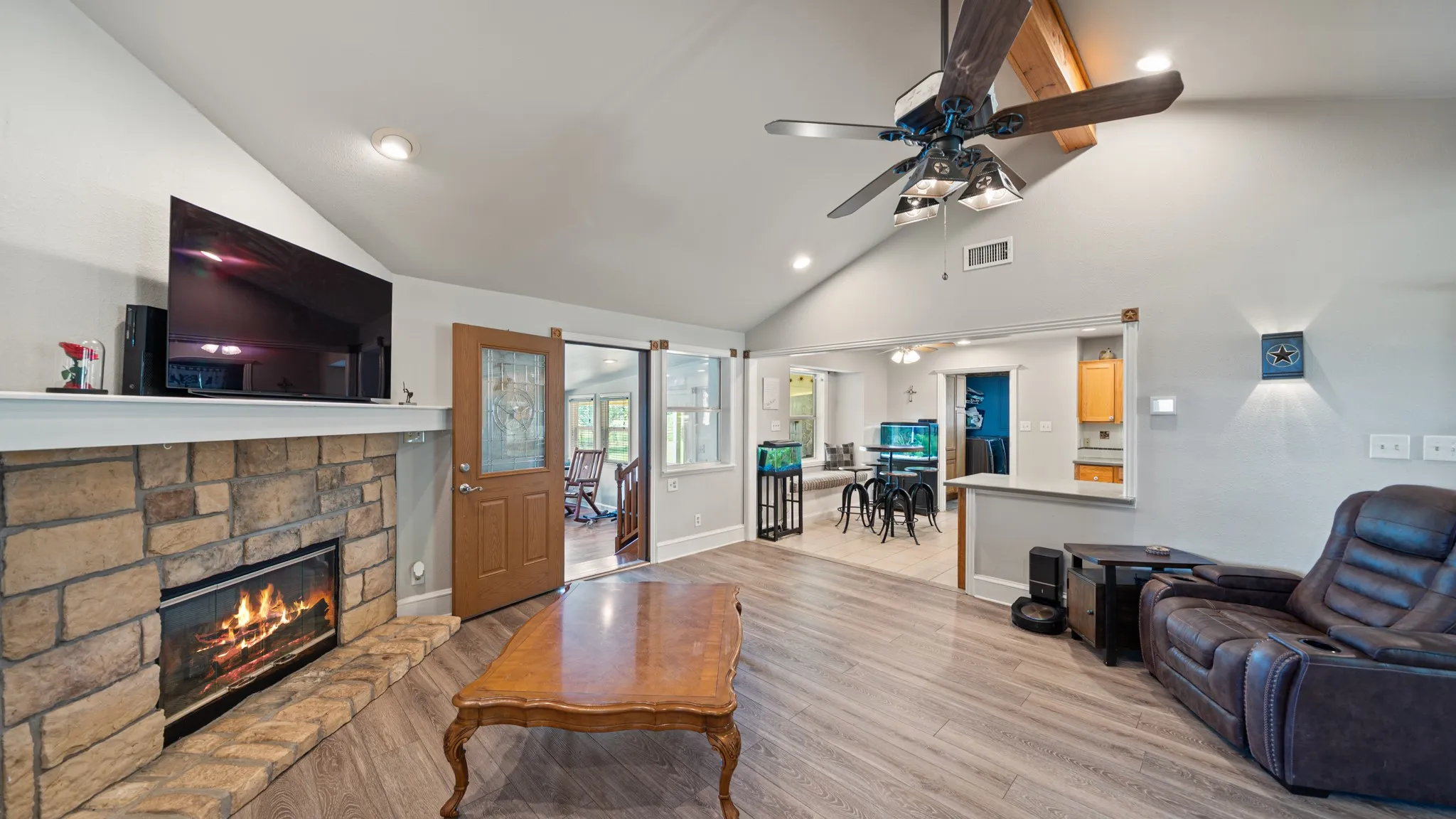 Open-concept living area featuring a wood-finish plank floor, ceiling fan with integrated lighting, and a gas fireplace with brick surround and mantel