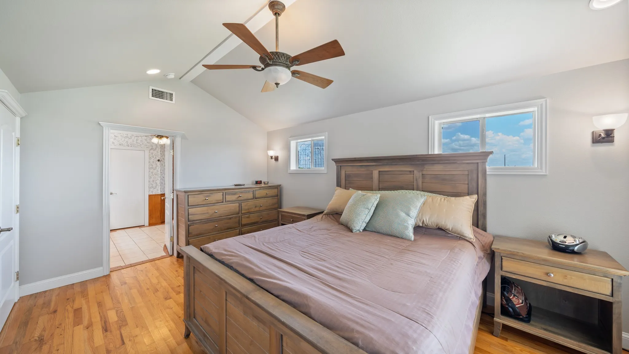 Master bedroom featuring wood-finish flooring, wood-constructed bed frame with upholstered headboard, dual nightstands with wood finish, ceiling fan with integrated lighting, and abundant natural light from multiple windows