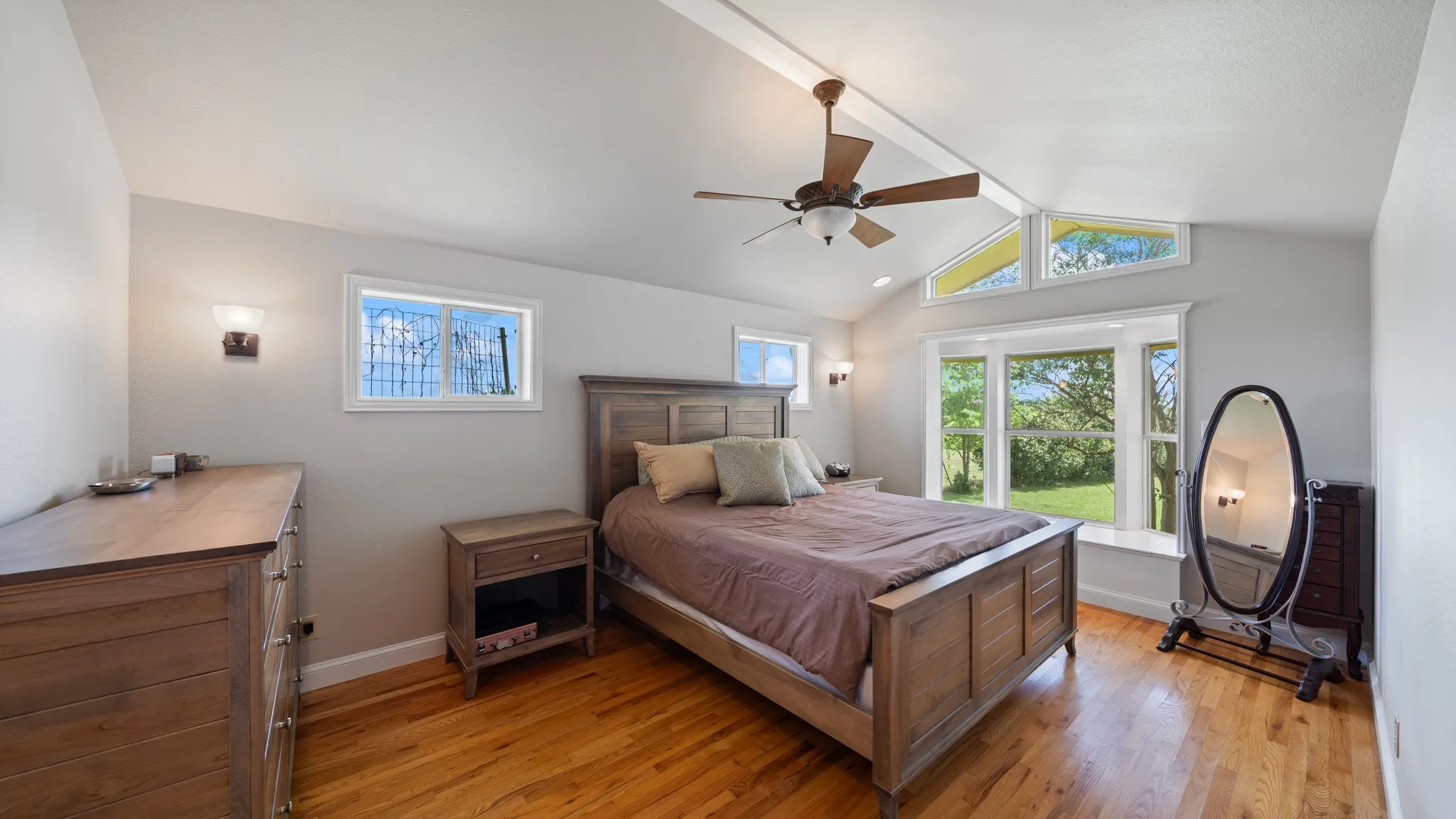 Bright primary bedroom showcasing hardwood flooring, wood-framed bed, ceiling fan with integrated lighting, and abundant natural light from multiple windows and glass doors