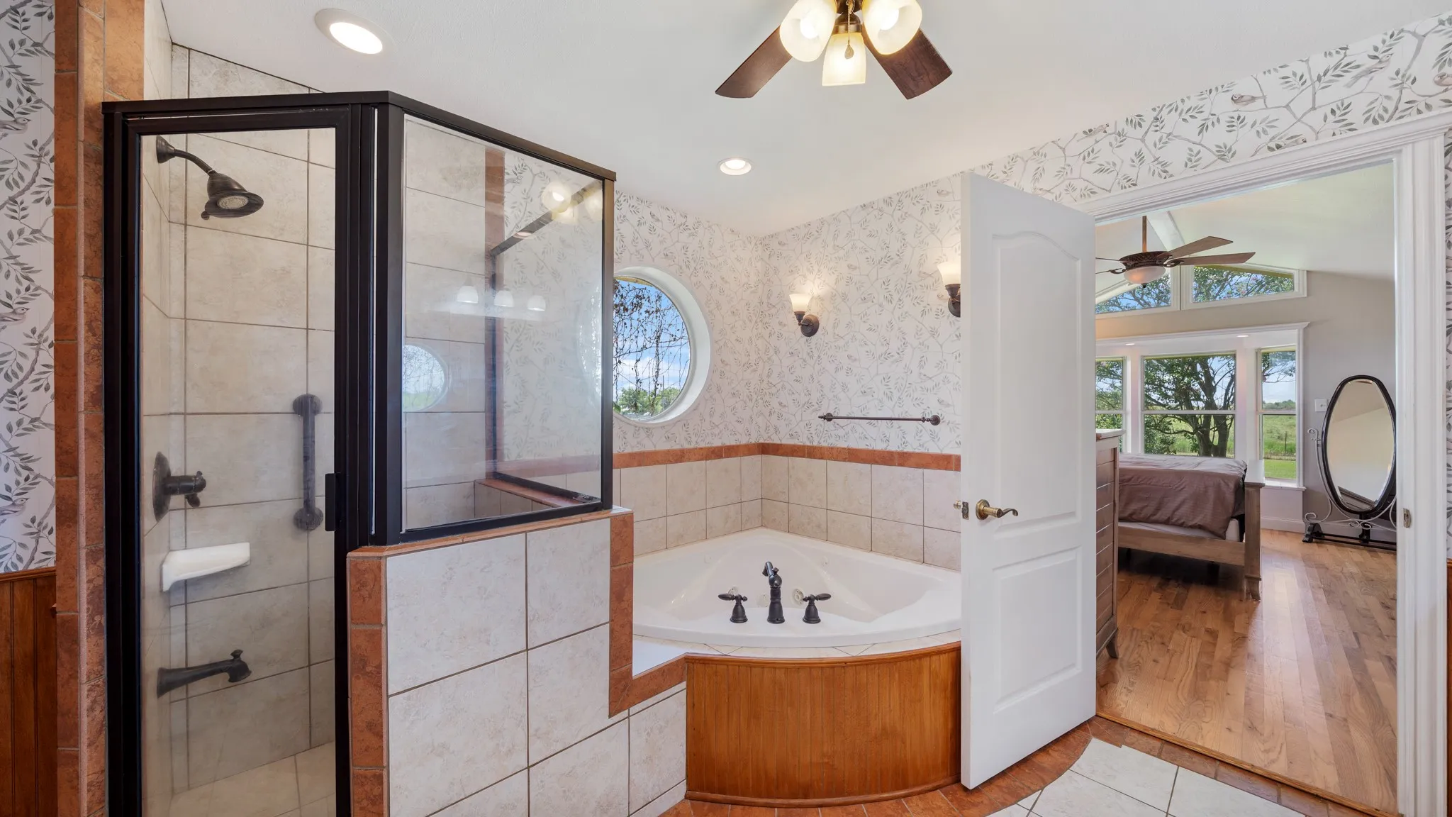 Luxurious primary bathroom featuring a corner soaking tub with wood-finish surround, a frameless glass-enclosed shower with tile surround, dual vanity with integrated storage, recessed ceiling lighting, and decorative tile wainscoting throughout
