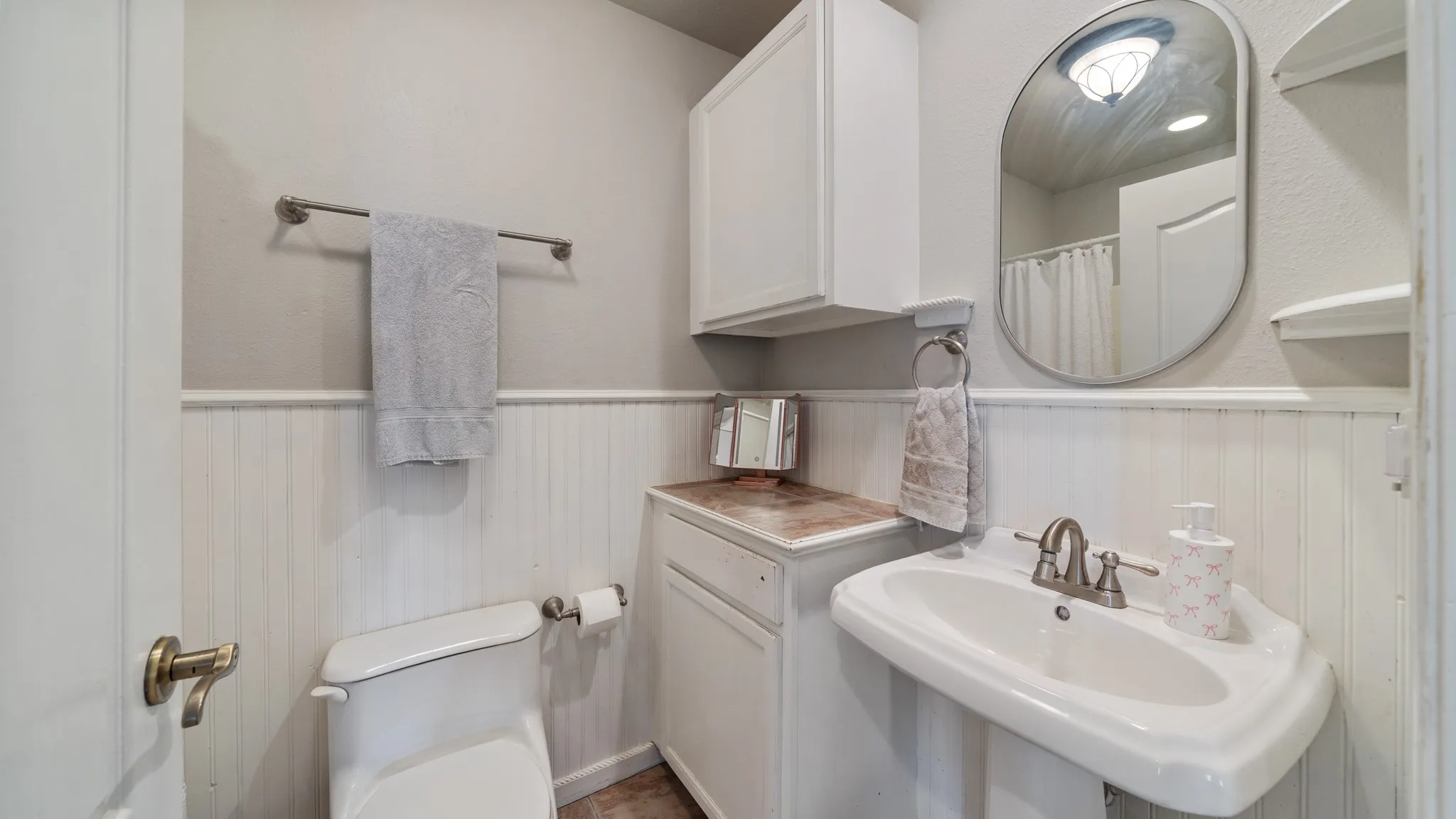 Bright bathroom featuring white subway tile wainscoting, pedestal sink with widespread faucet, white cabinetry with wood-finish countertop, arched mirror with integrated shelving, and hardwood flooring