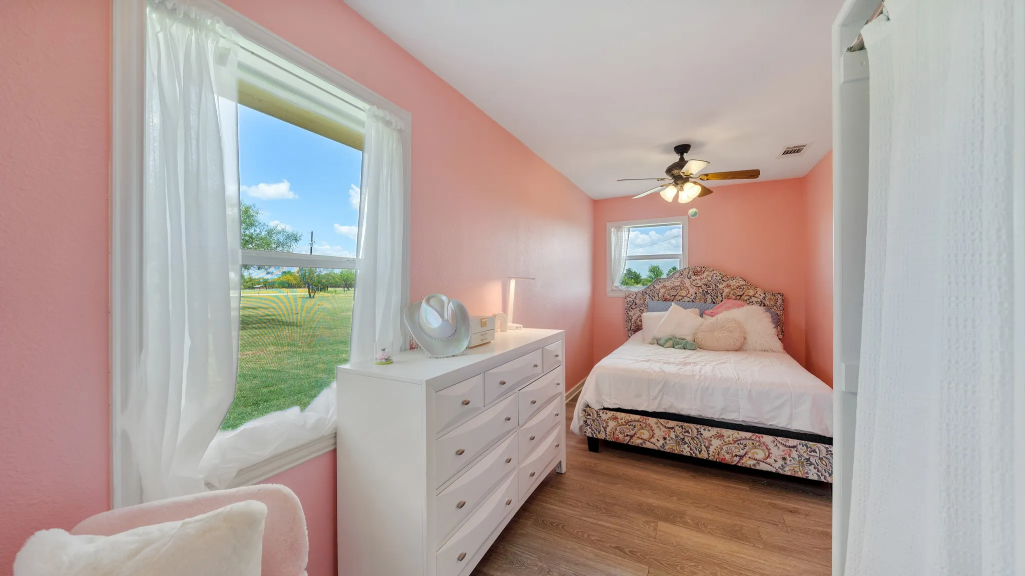Charming bedroom with coral-hued walls, wood-finish flooring, and abundant natural light from a large window framed with sheer curtains