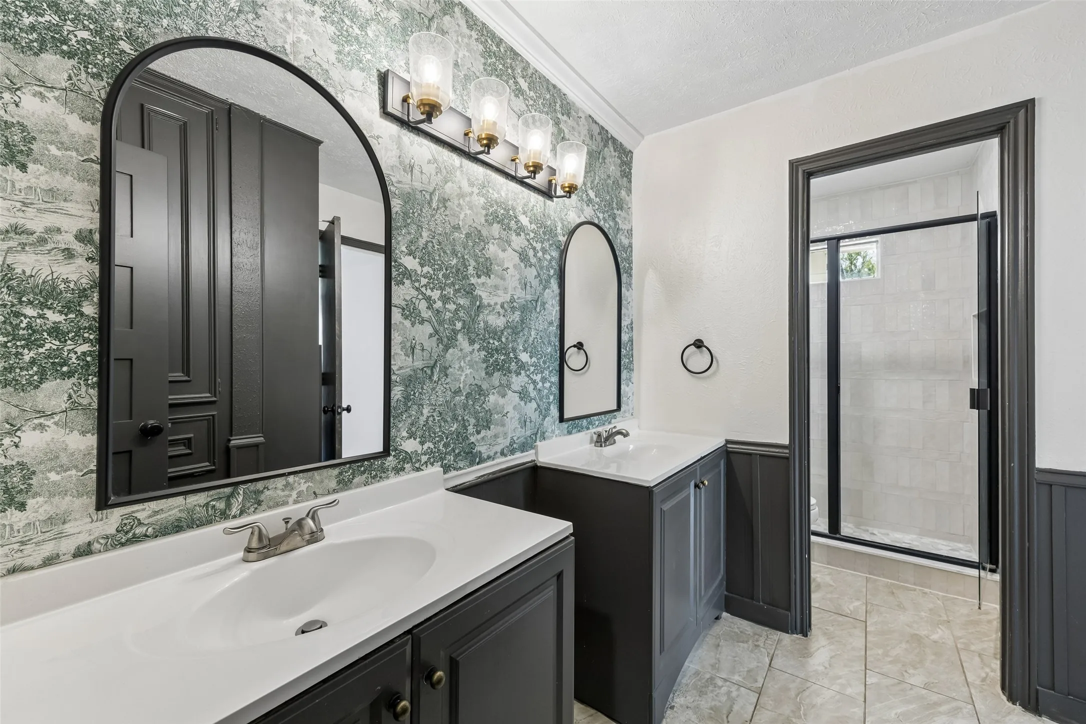 Dual vanity with undermount sinks and dark cabinetry, arched mirrors with dark frames, patterned wallcovering, and a shower with a framed glass enclosure