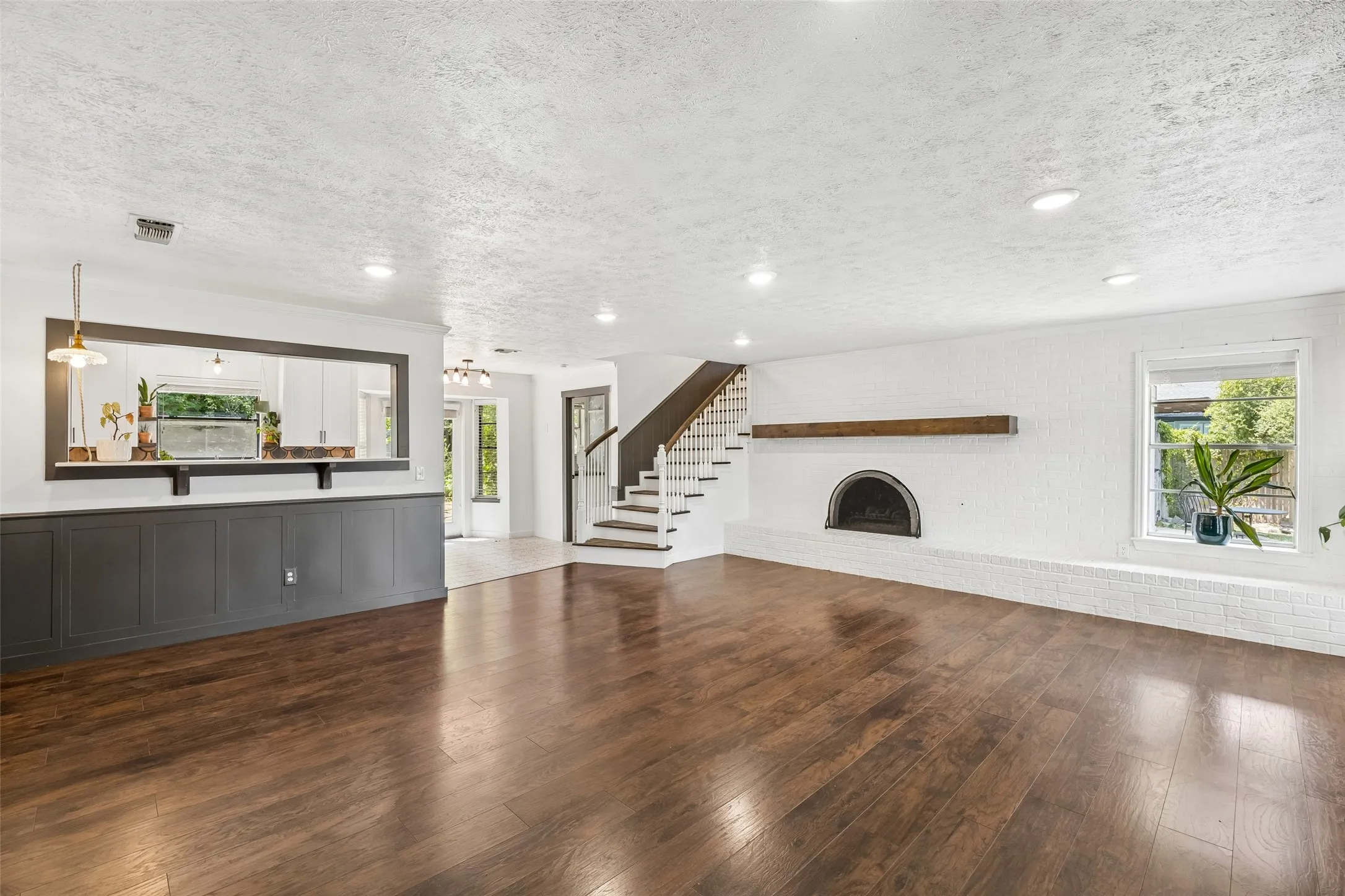 Wood-finish flooring throughout the room, white brick fireplace with dark arched opening, dark wood mantel, recessed lighting, and a white framed window