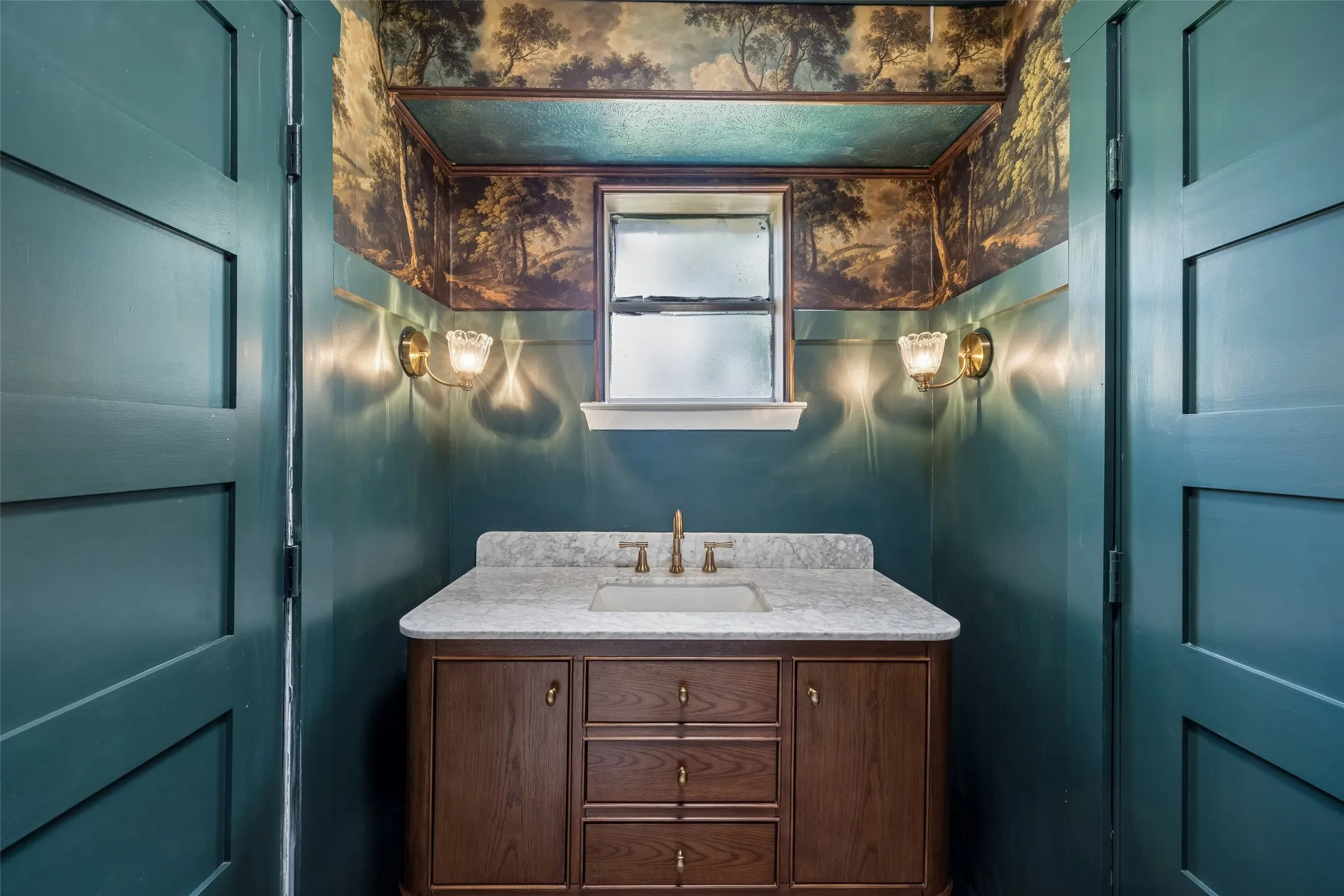 Full bath featuring, a wood vanity with marble-top countertop and brass hardware, dual brass sconces with milk glass shades, and a central window with decorative wallpaper
