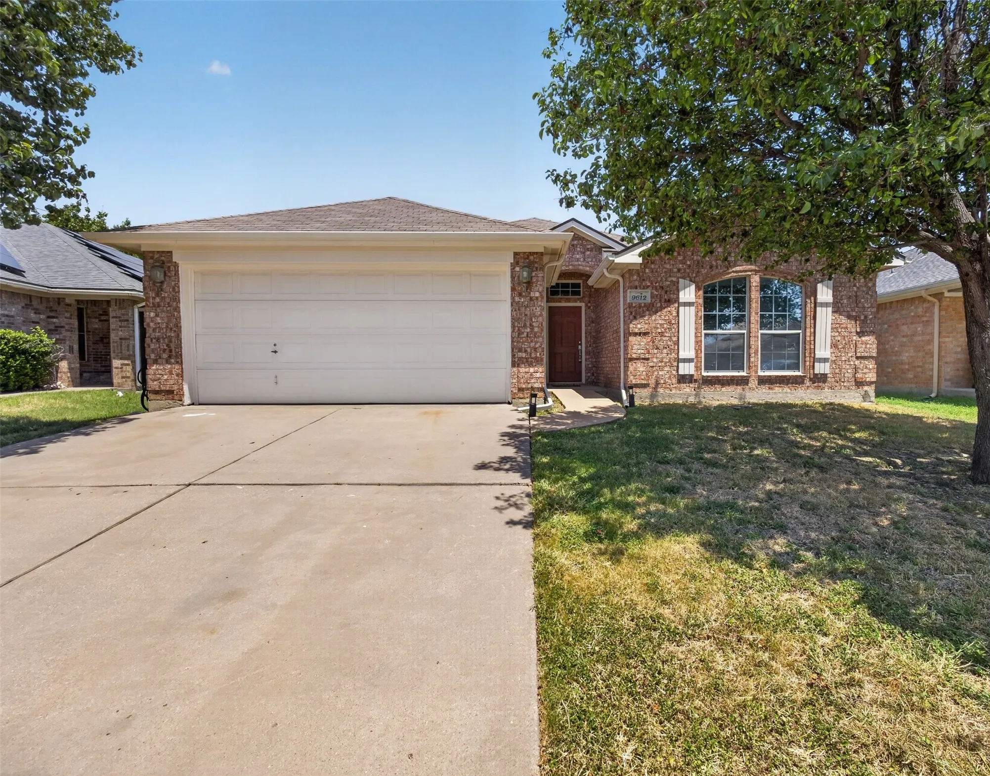 Welcome home to this charming one-story brick residence featuring a spacious two-car garage with white panel doors, a wide concrete driveway, and a manicured front lawn shaded by mature trees