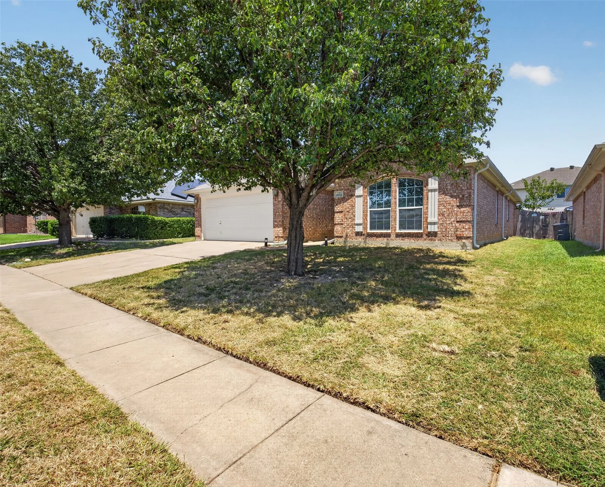 Brick exterior with a white garage door, featuring decorative window shutters, a concrete driveway, and mature trees