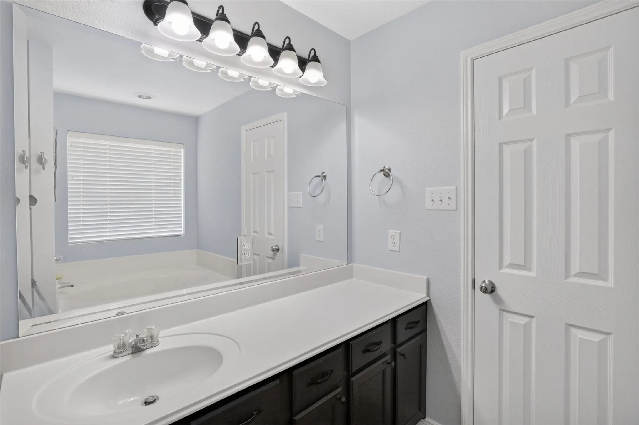 Spa-inspired master bathroom featuring a generous dual-sink vanity with black cabinetry and white countertop, large framed mirror with integrated lighting, and separate soaking tub