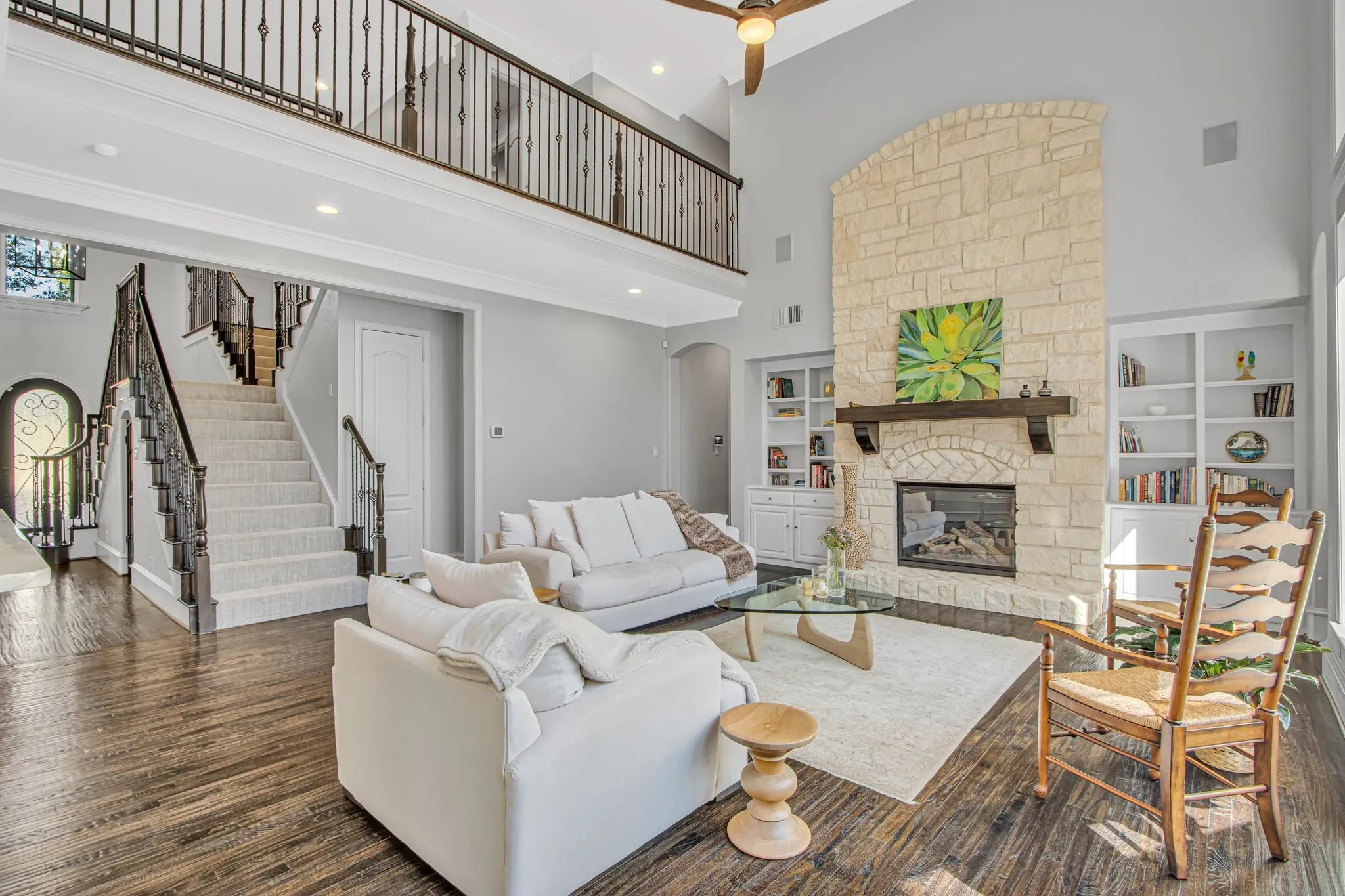 Soaring two-story living room anchored by a stacked-stone fireplace with built-in shelving, hardwood flooring throughout, and abundant natural light from high ceilings.