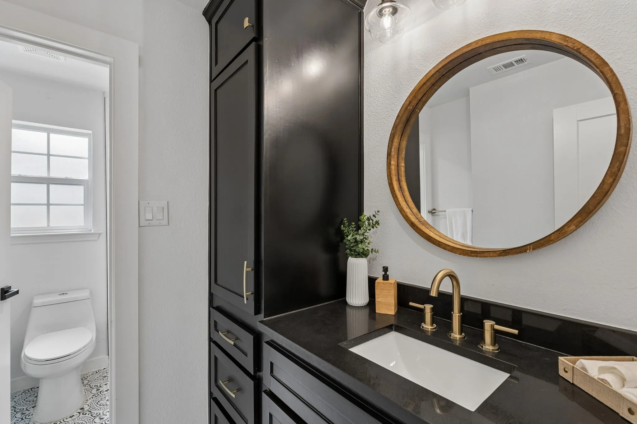 Built-in dark cabinetry with gold-tone hardware, dark countertop with an undermount sink, gold-tone faucet, and a round wall mirror