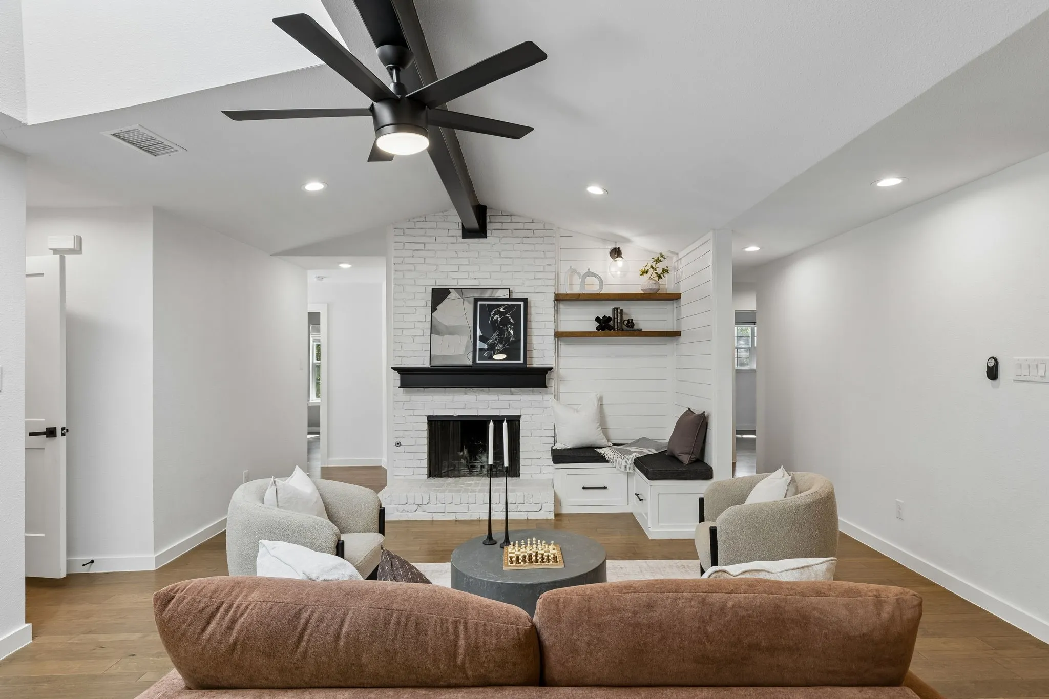 Vaulted ceiling with exposed beams and a ceiling fan, brick-faced fireplace with a black mantel, built-in window seating with storage, wood-finish flooring, recessed lighting throughout