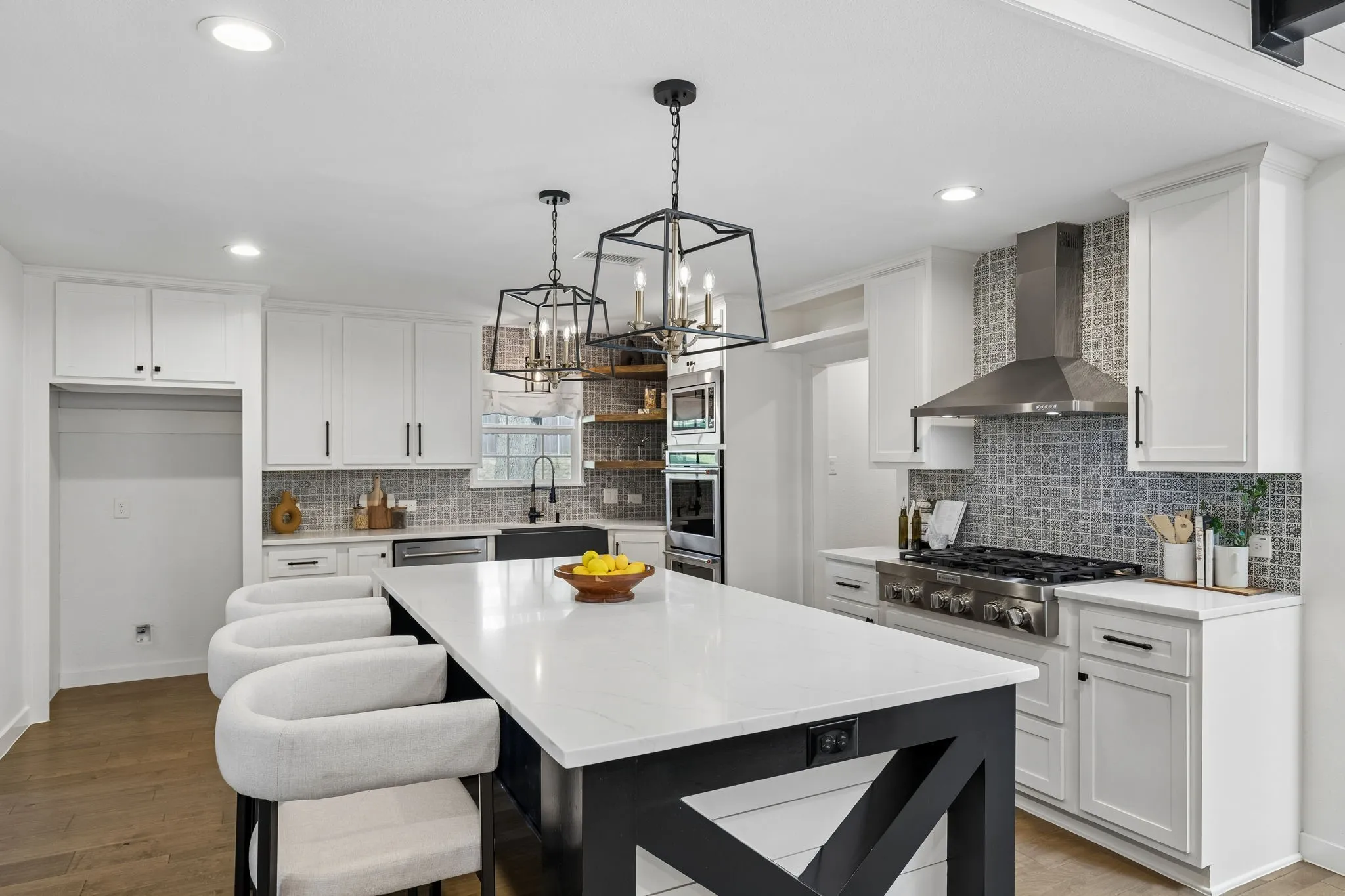 White cabinetry with black hardware, wood-finish flooring, patterned tile backsplash, stainless steel range hood, and a kitchen island with a light-toned countertop