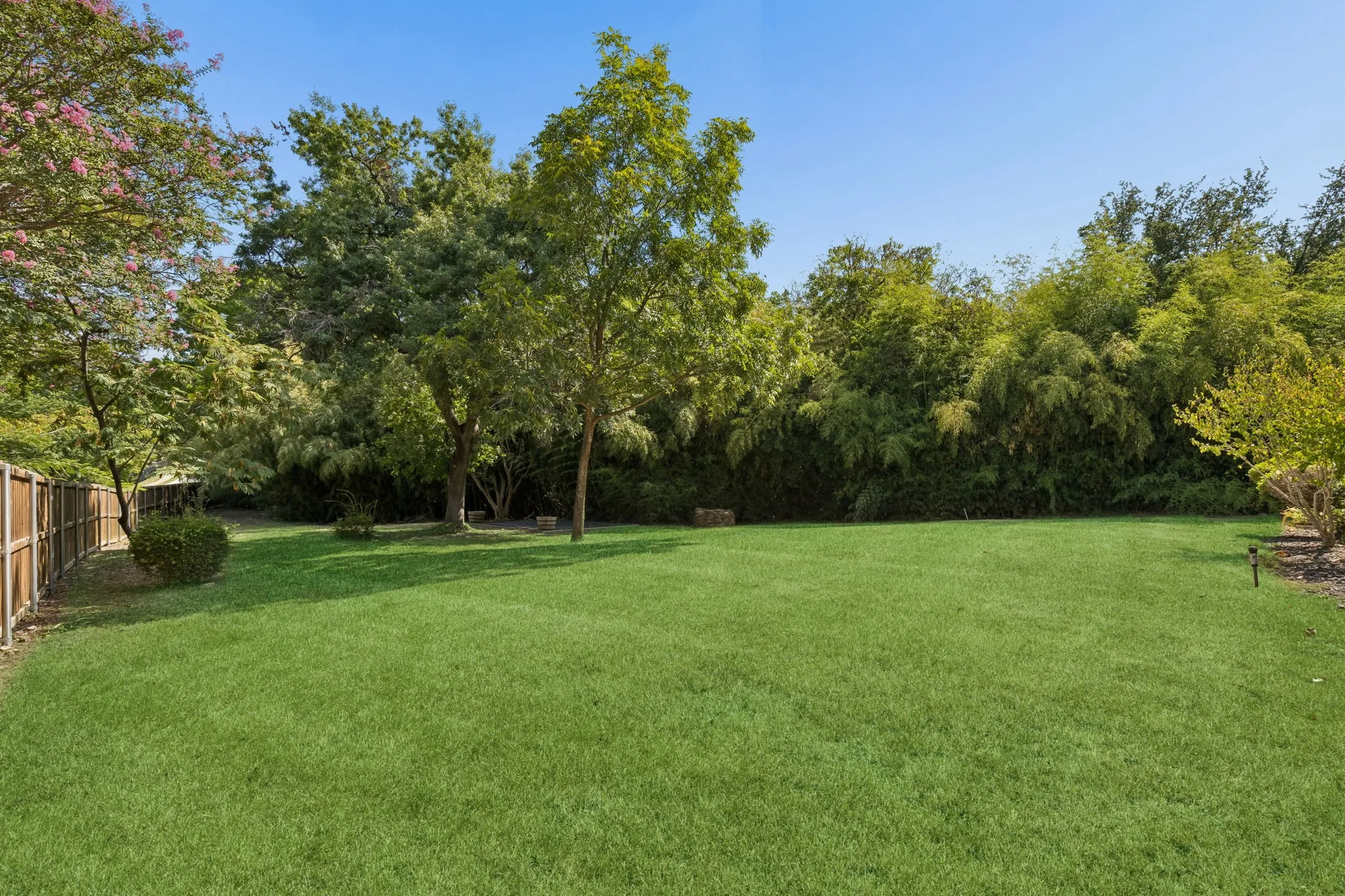 Expansive lawn area bordered by mature trees and shrubs, featuring a wood fence, and landscape lighting fixtures