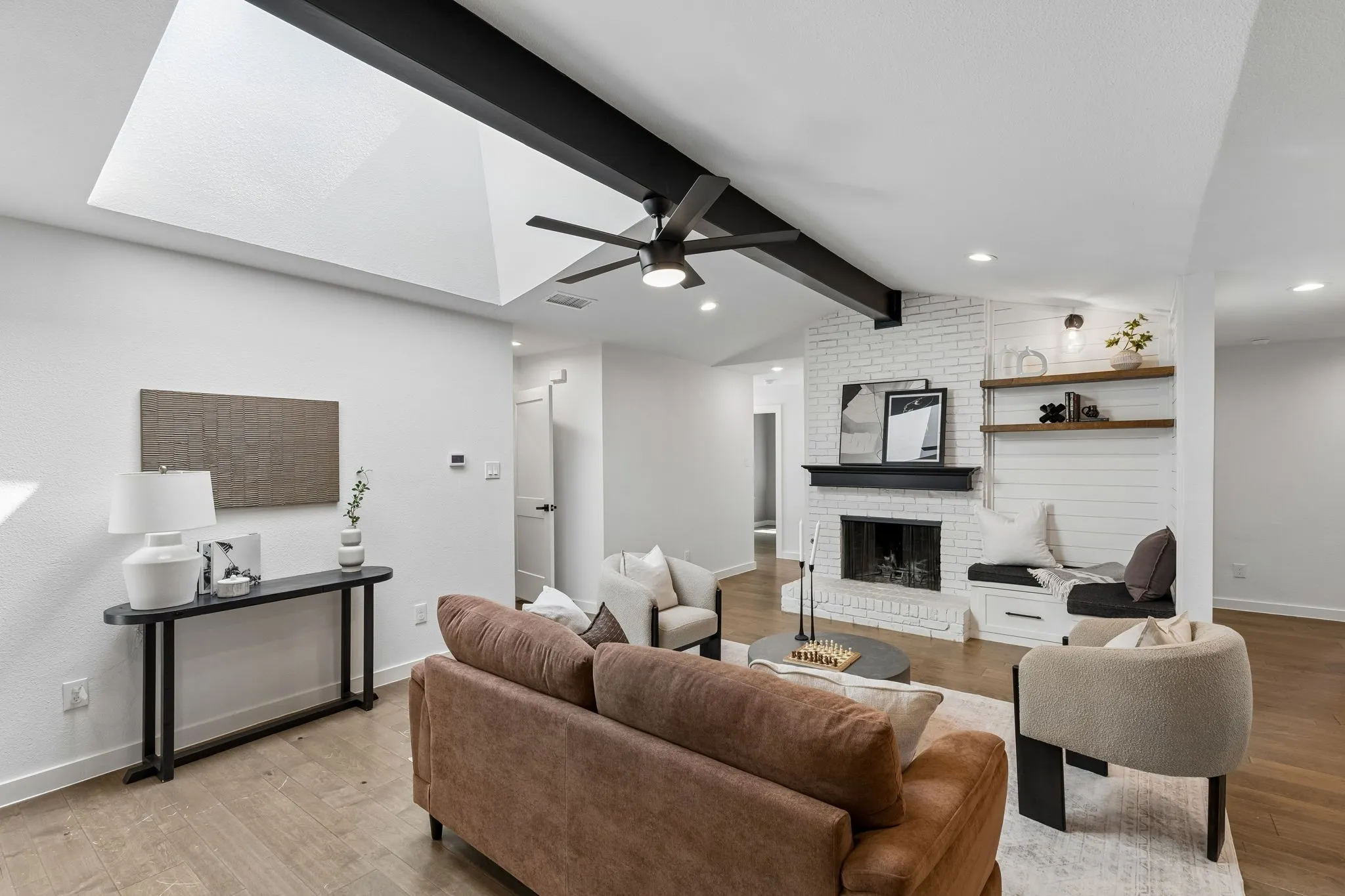 Vaulted ceiling with skylight and dark exposed beams, white painted brick fireplace with a dark mantel, wood-finish flooring, recessed lighting, and a built-in bench with shelving