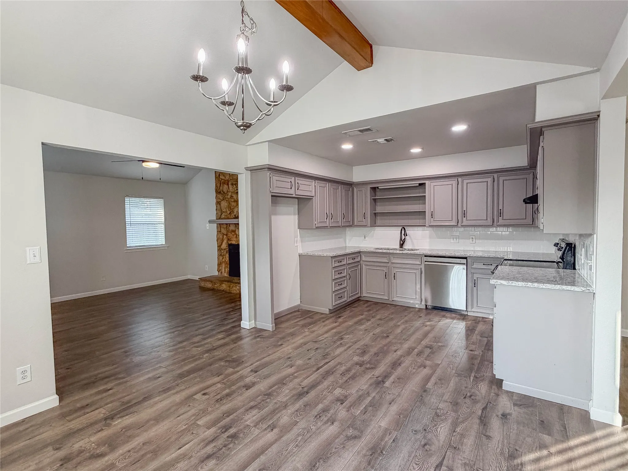 Vaulted ceiling with wood beam accent, wood-finish flooring, stone fireplace, updated kitchen cabinetry, and stainless steel appliances