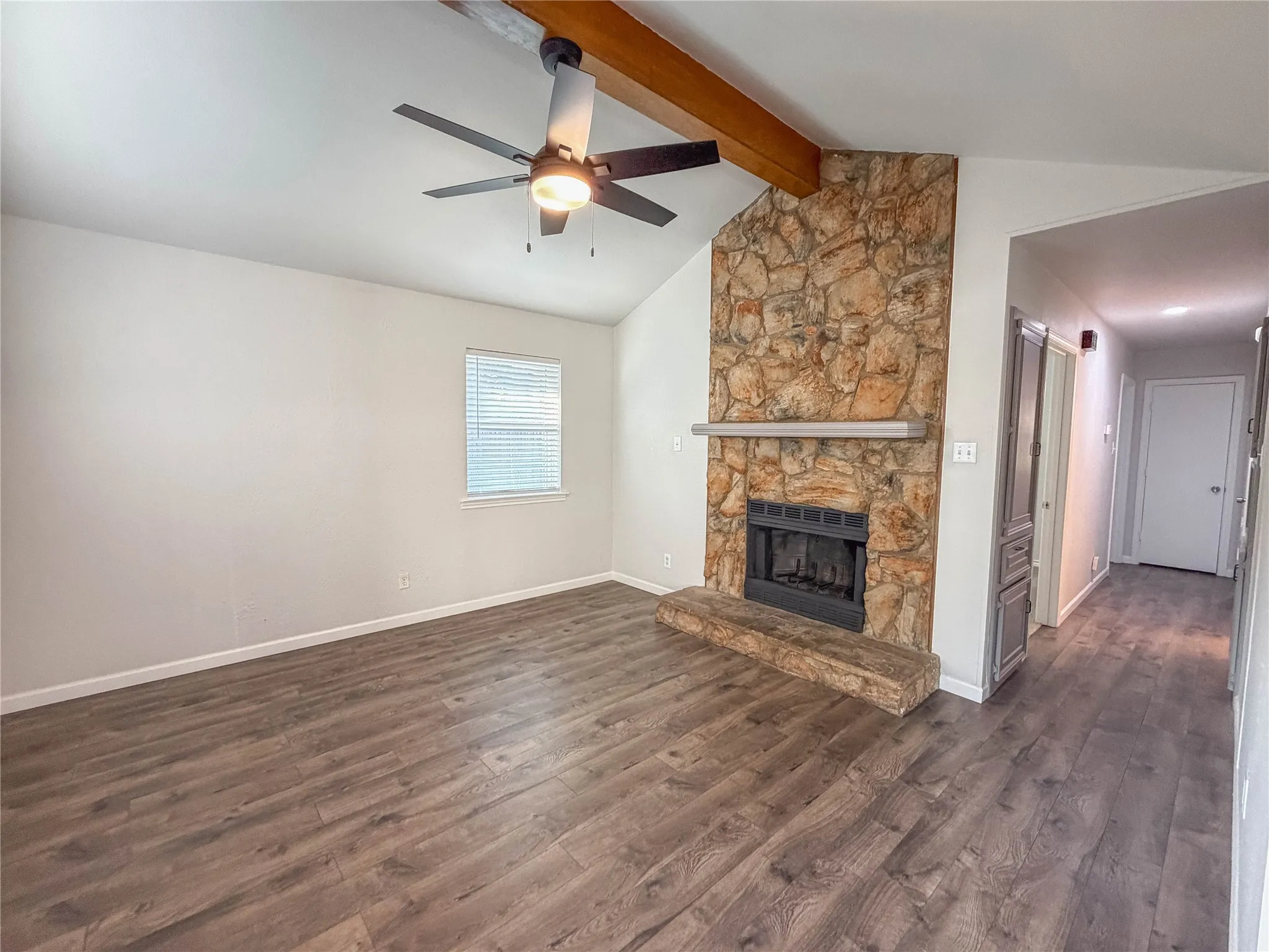 Vaulted ceiling with exposed wood beam, stone-clad fireplace with mantel, wood-finish flooring, and ceiling fan with integrated lighting