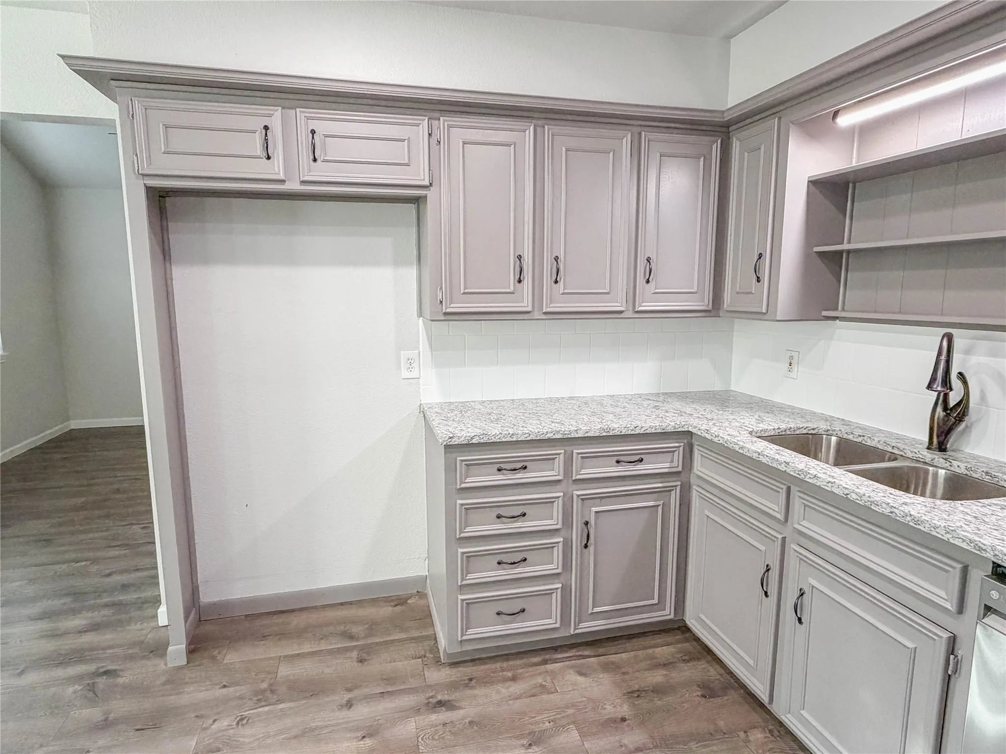 Light gray cabinetry with crown molding, granite-style countertops, white subway tile backsplash, dual stainless steel sink, and wood-finish flooring