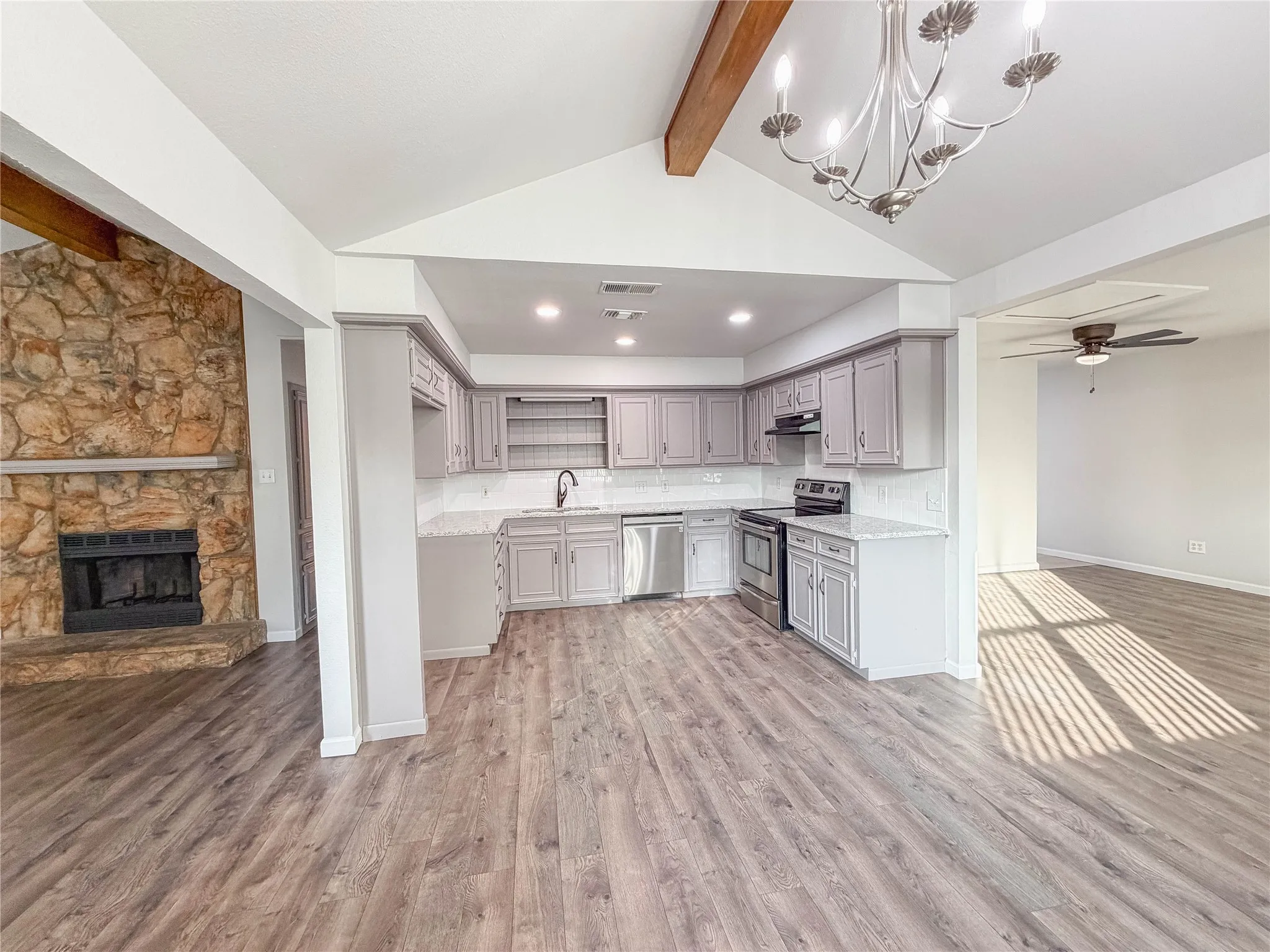 Vaulted ceiling with exposed wood beam, kitchen with gray cabinetry and stainless steel appliances, stone-faced fireplace with hearth, and wood-finish flooring