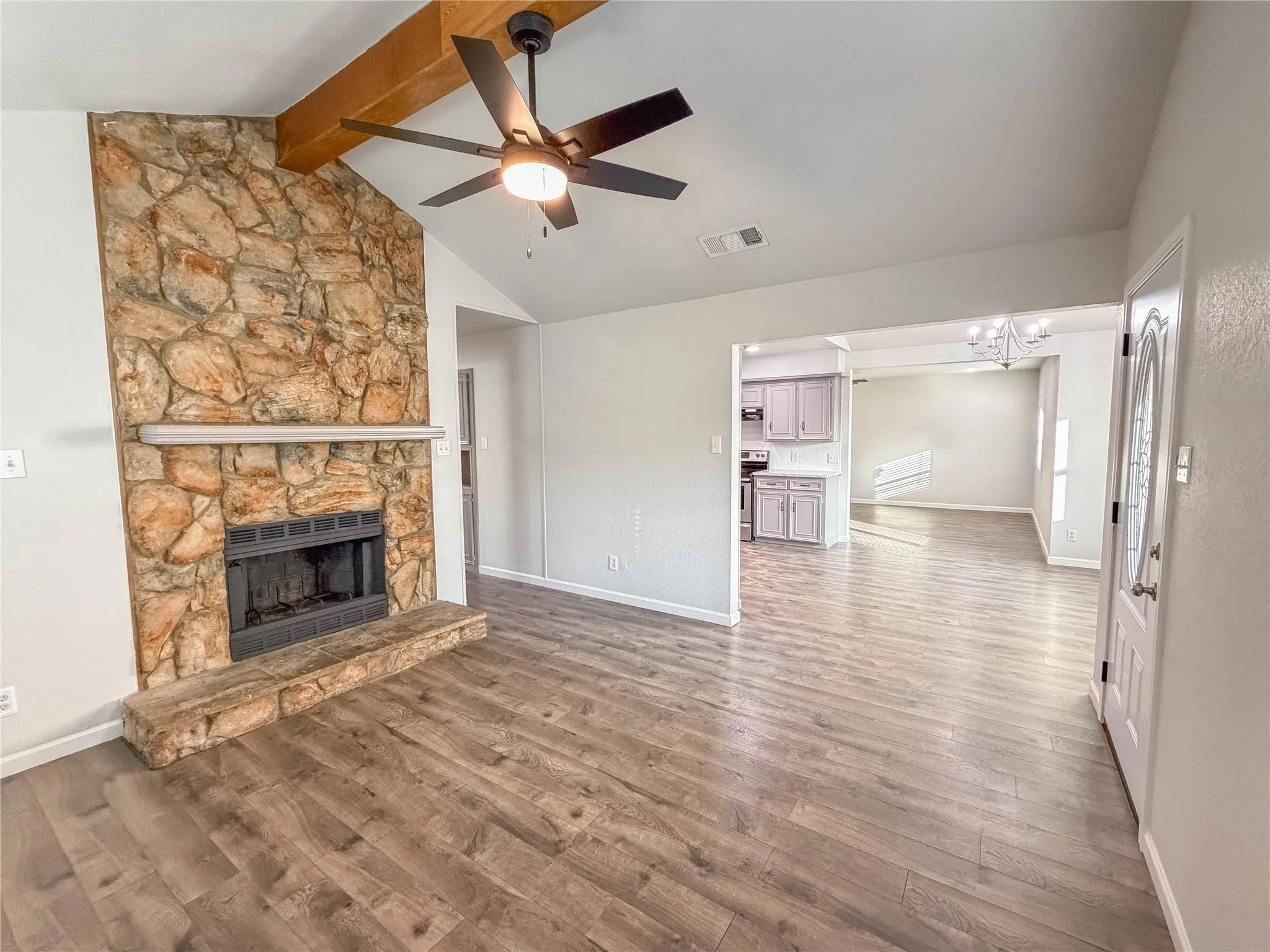 Vaulted ceiling with exposed wood beam, stone-clad fireplace with hearth, wood-finish flooring, and recessed kitchen beyond