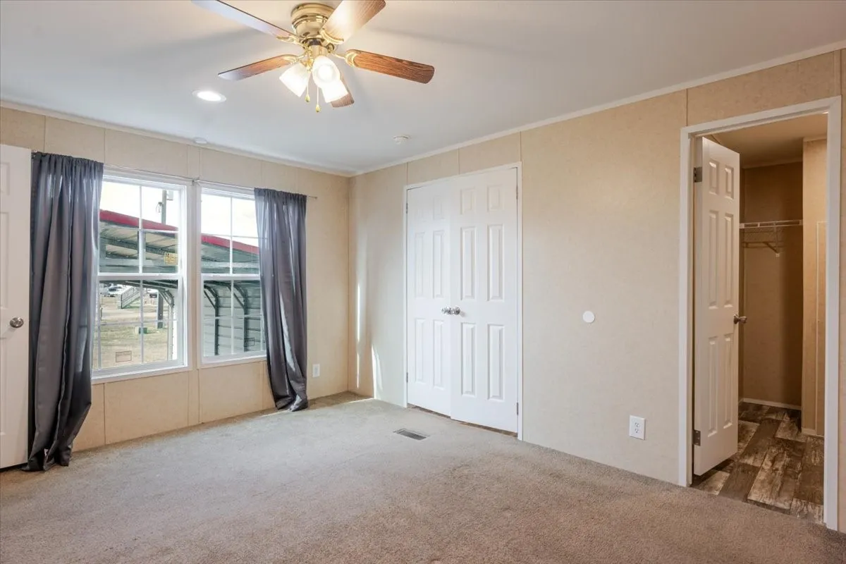 Carpeted interior room featuring a ceiling fan with light fixture, two windows with curtains, a built-in closet with bifold doors, and an additional open closet with wood-finish flooring
