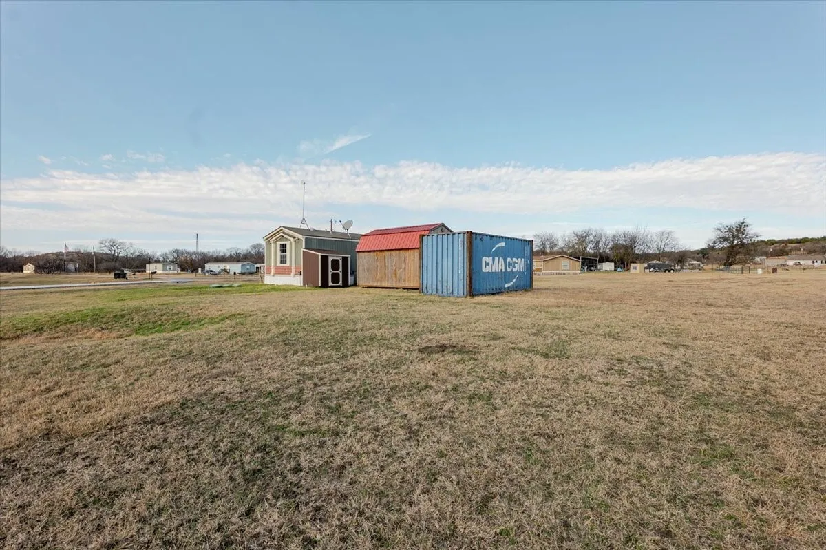 Open field with a small structure featuring light-colored siding and a dark roof, accompanied by a blue shipping container and a red-roofed shed