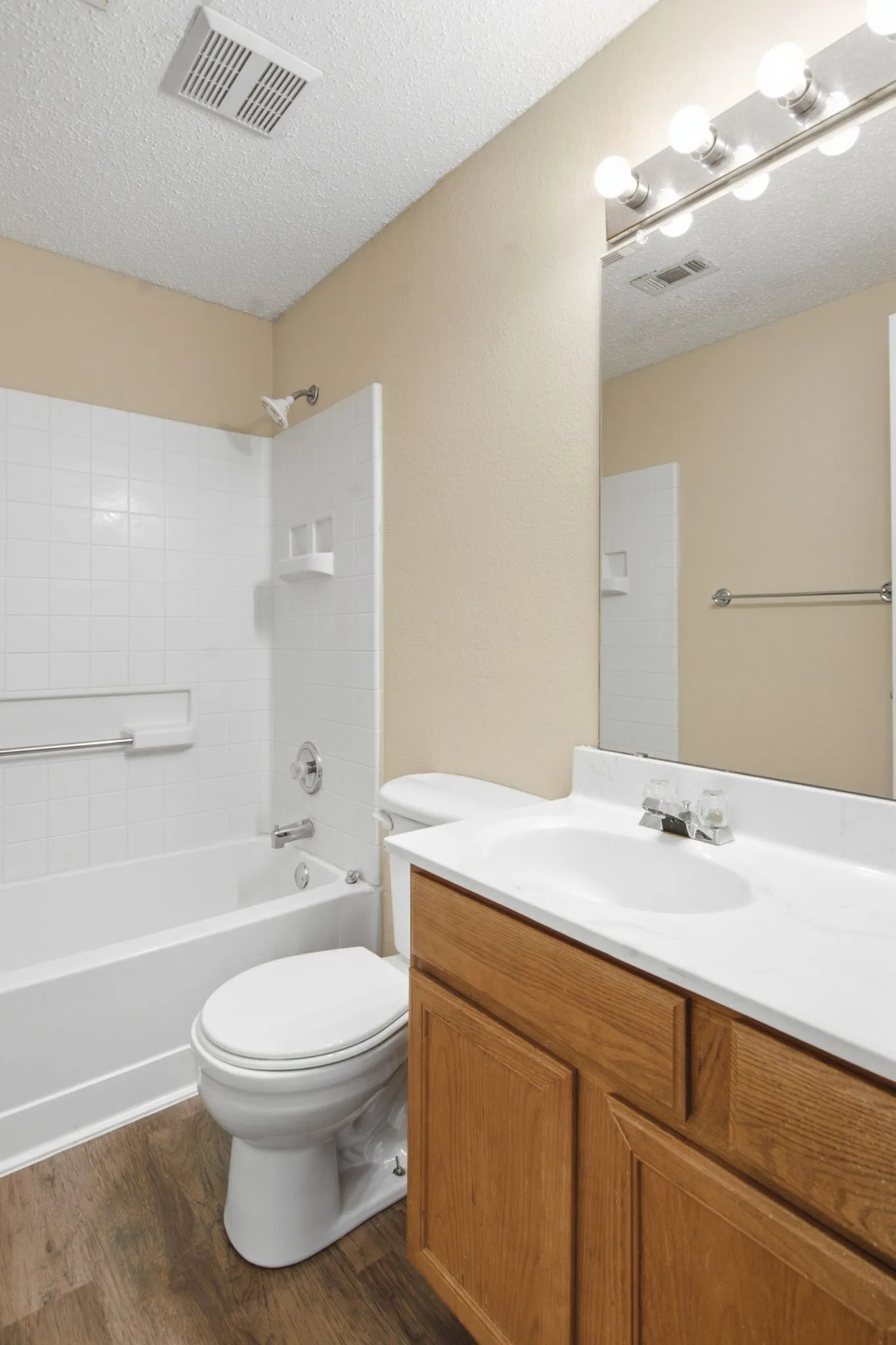 Wood-finish flooring, white bathtub with tiled surround, wood vanity with integrated sink, chrome hardware, and mirror with overhead vanity lighting