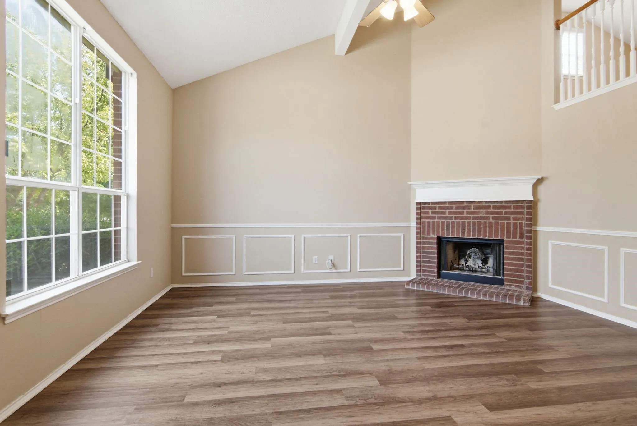 Vaulted ceiling with exposed beam, wood-finish flooring, large window with grid pattern, brick fireplace with mantel, and decorative wainscoting