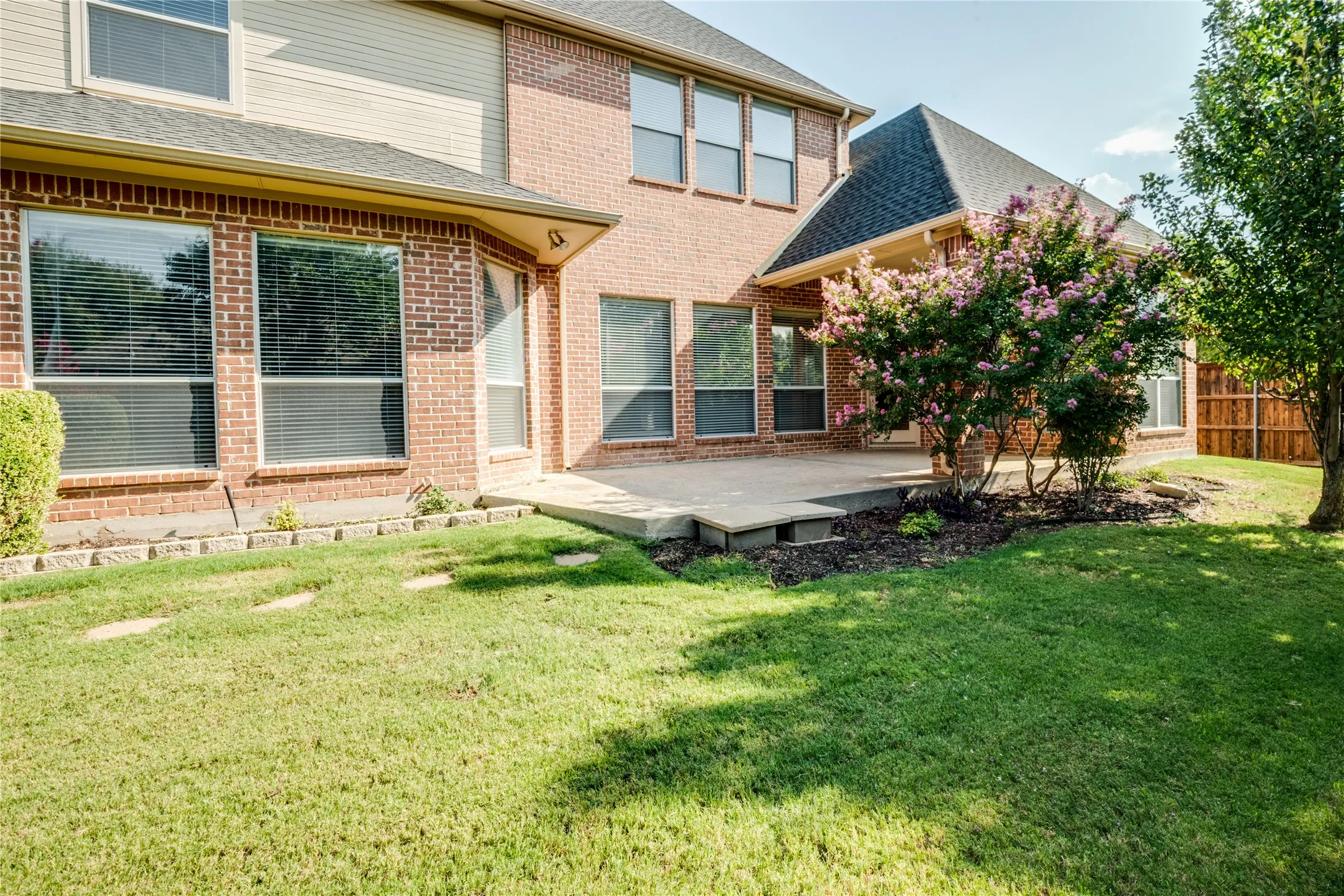 Rear exterior featuring brick and siding construction, a concrete patio, a mature flowering shrub, and a wooden privacy fence