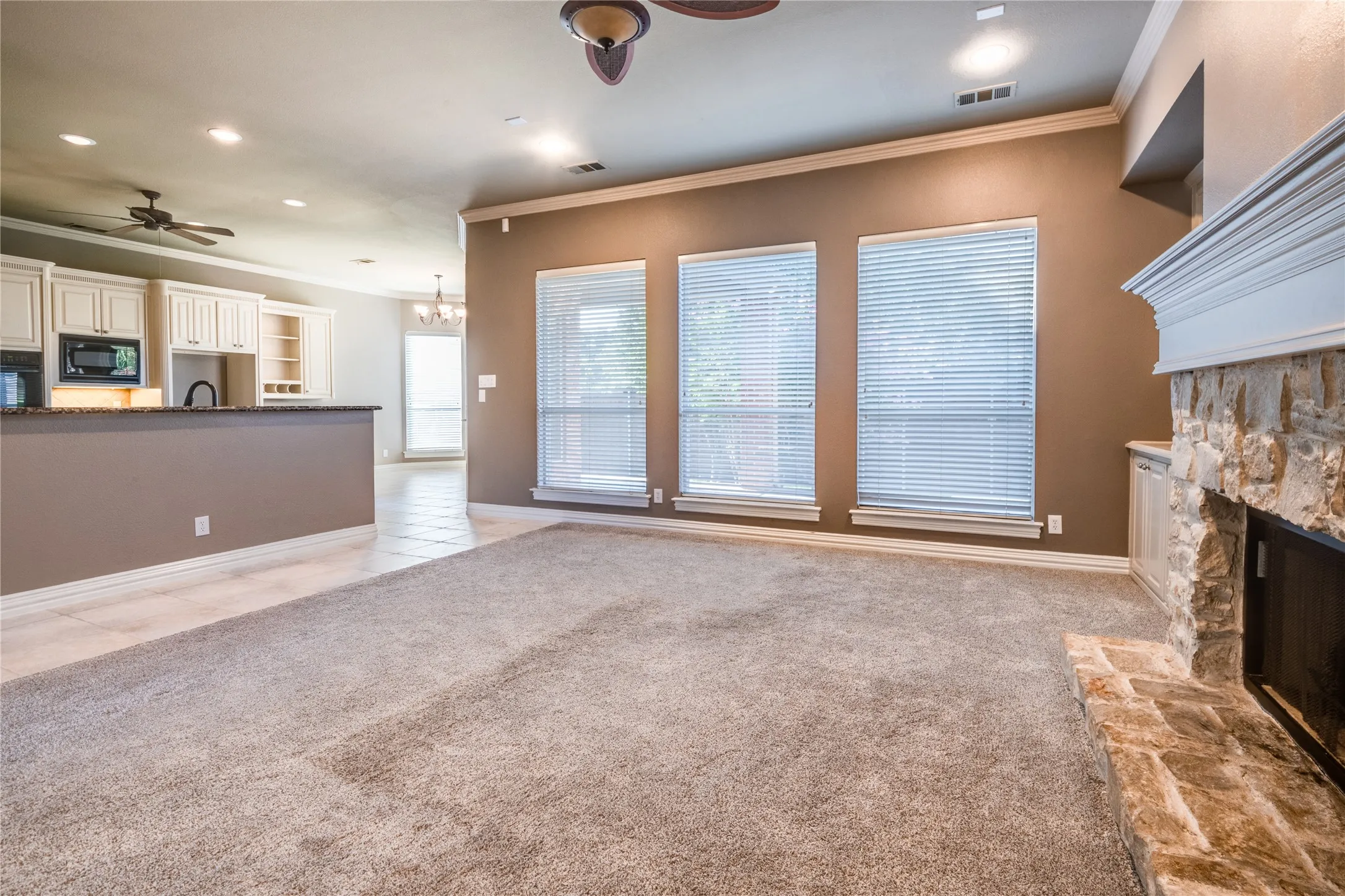 Open-concept living area with a stone fireplace, three large windows, crown molding, and recessed lighting