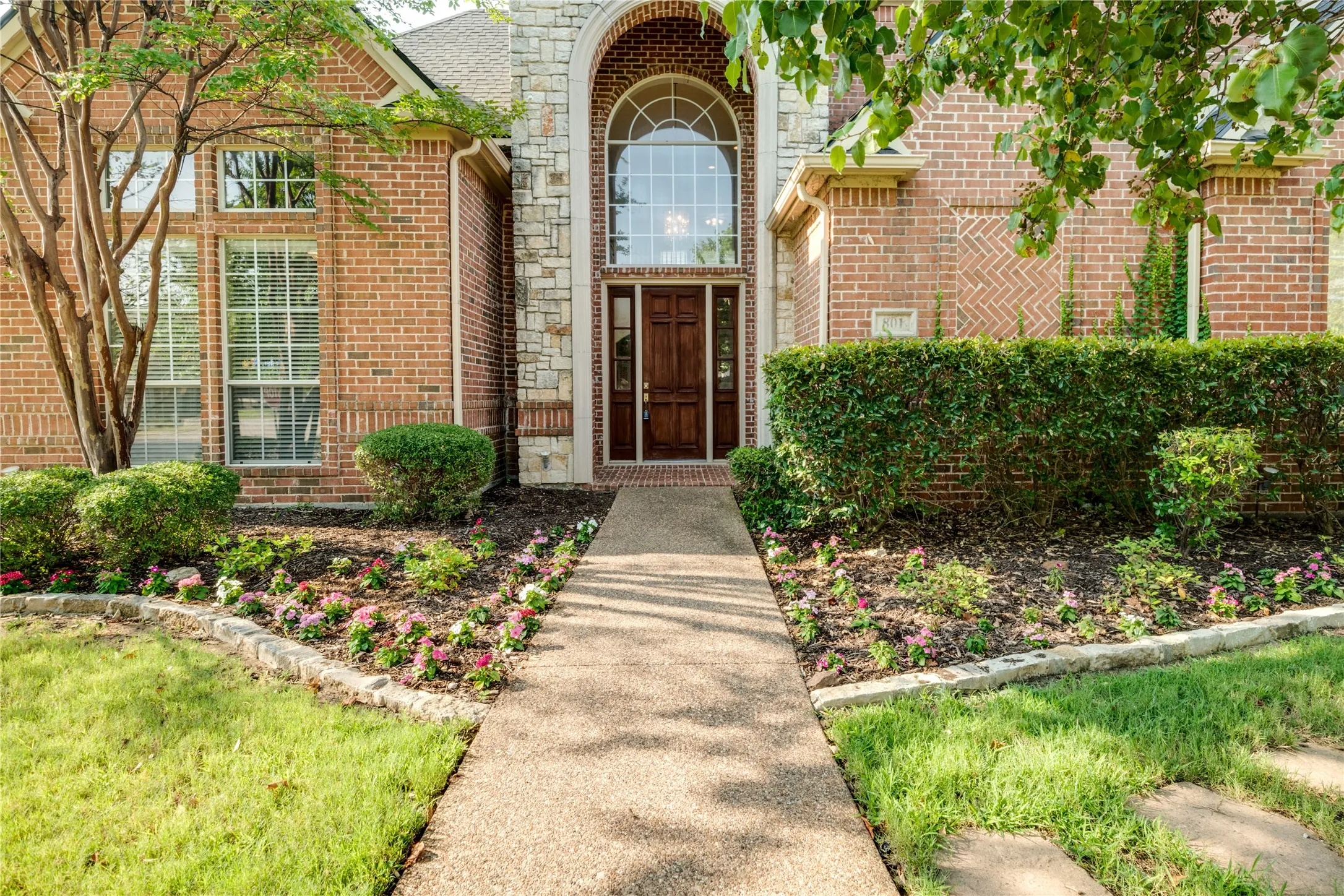 Brick facade with stone accents, arched entry with transom window, wood-paneled front door, and manicured landscaping