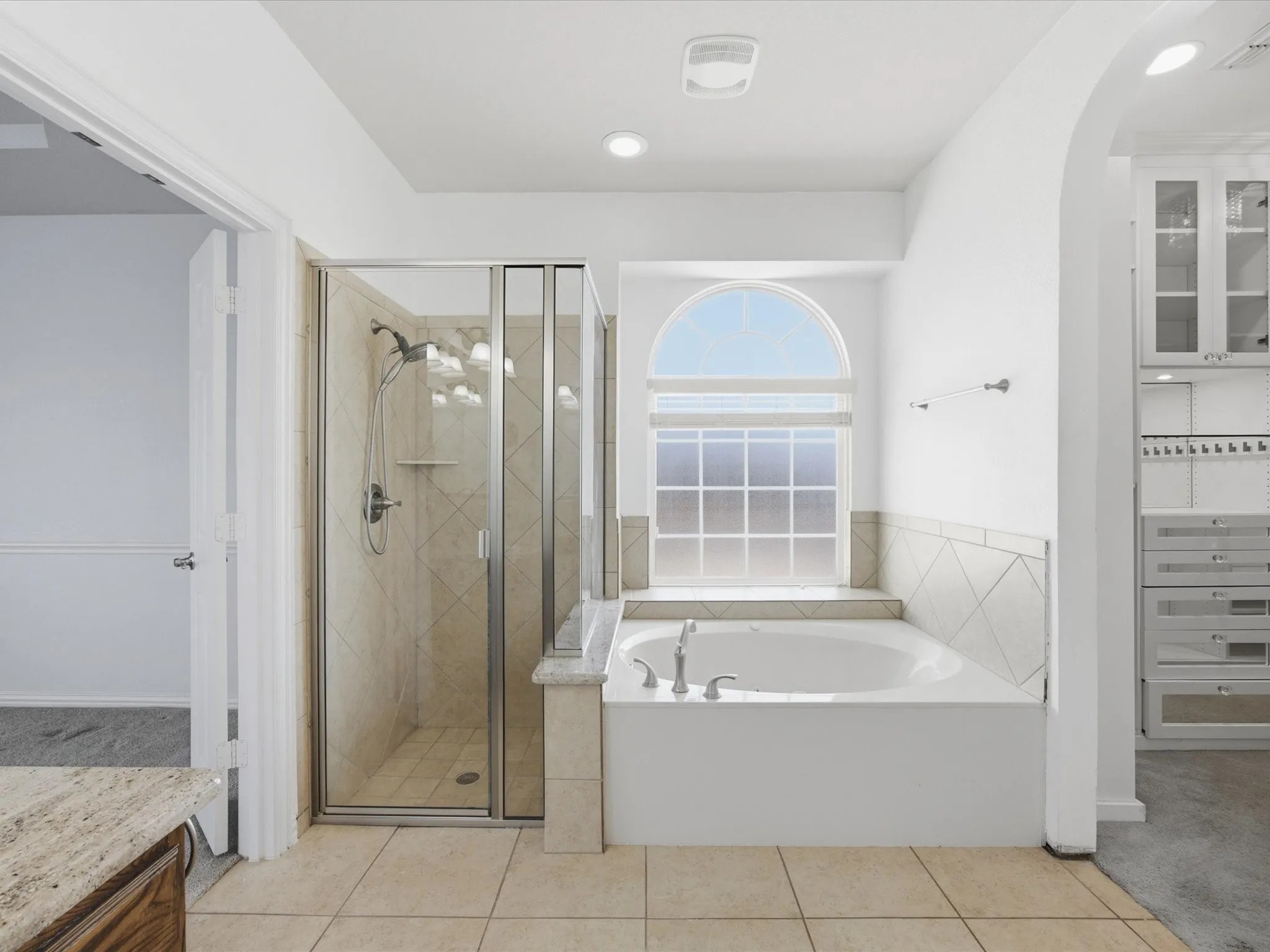 Enclosed glass shower with tiled surround, soaking tub with arched window, and custom built-in cabinetry