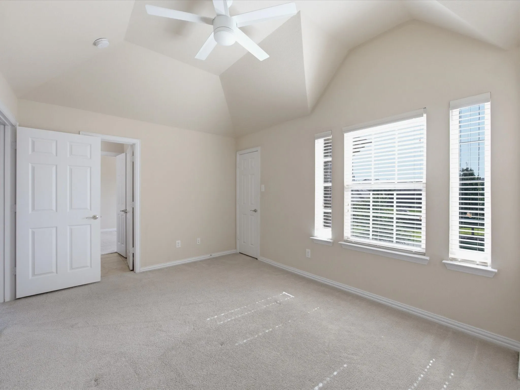 Bright, light-filled bedroom with vaulted ceiling and ceiling fan, abundant natural light from large multi-pane windows with plantation shutters, neutral carpet flooring, and double closet storage