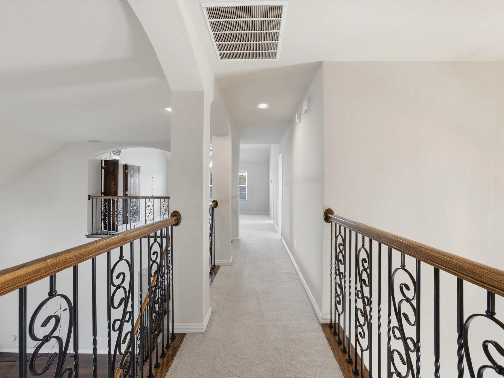 Carpeted hallway with arched openings, recessed lighting, and ornate wrought iron and wood-finish railings