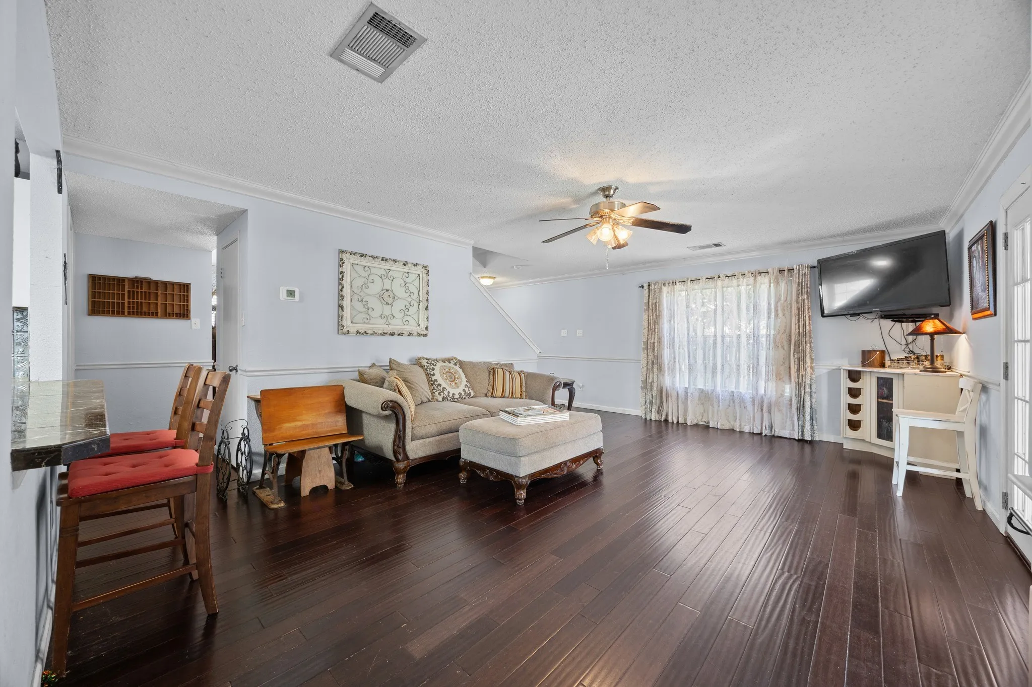 Spacious living area showcasing dark wood-finish flooring, a ceiling fan with integrated lighting, and large windows with sheer curtains