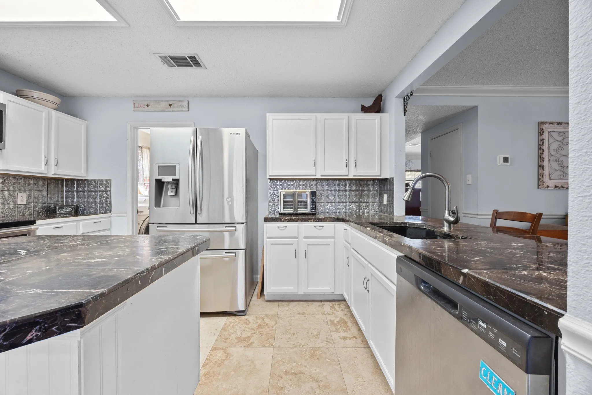 Modern kitchen featuring white cabinetry, stainless steel appliances including a French-door refrigerator and dishwasher, dark stone countertops with waterfall edges, subway tile backsplash, recessed ceiling lighting, and tile flooring throughout the open layout