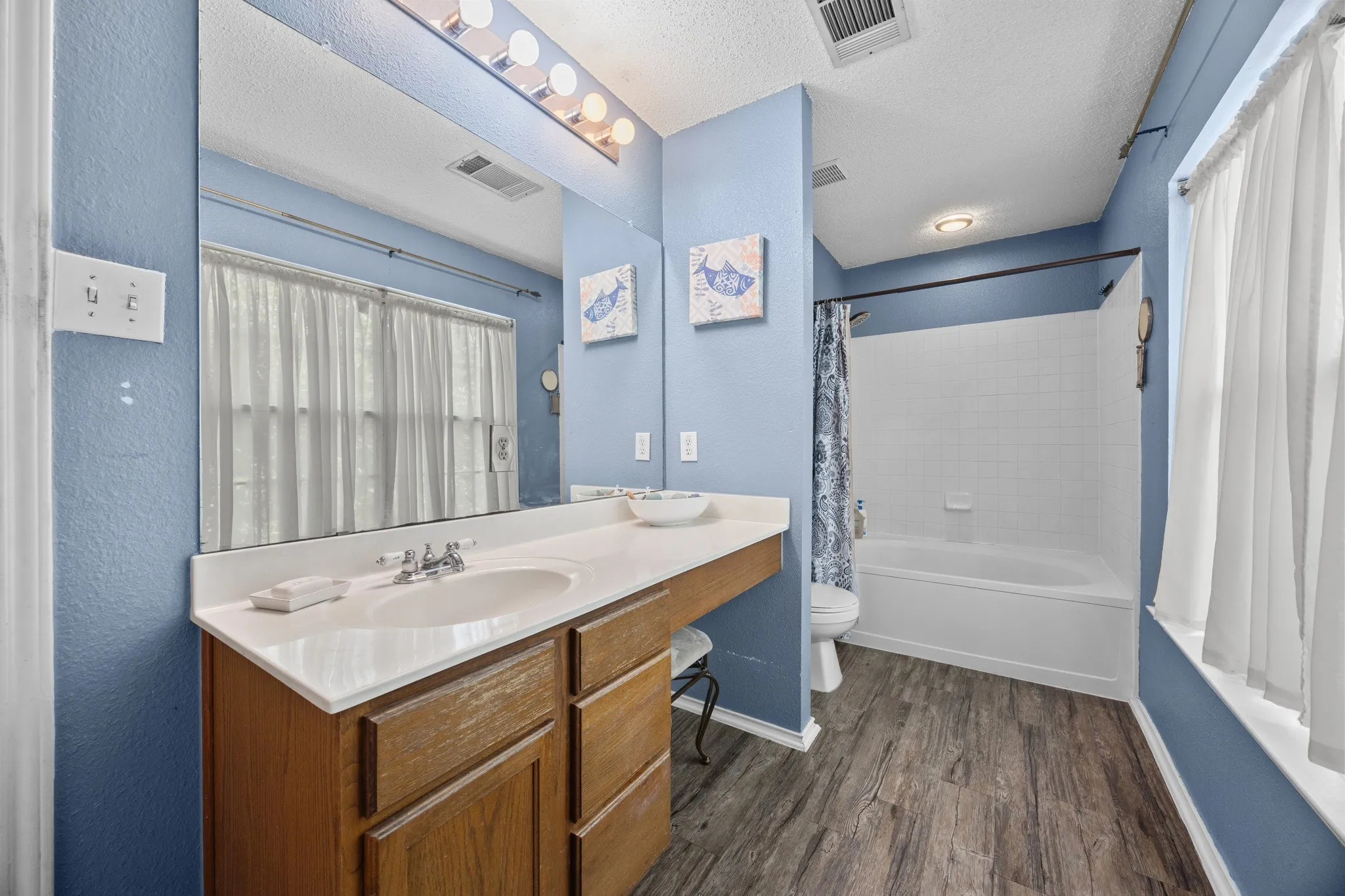 Single vanity with a white countertop and wood-finish cabinetry, large wall-mounted mirror, white bathtub with a tiled surround, and wood-finish flooring