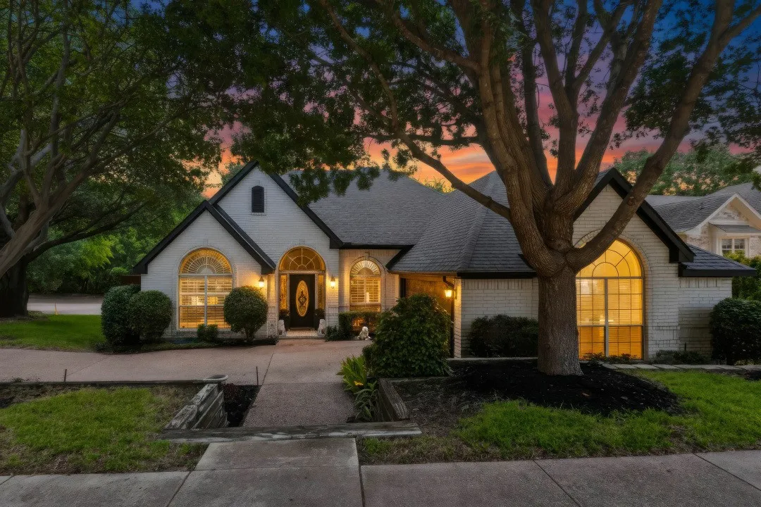 White brick exterior with dark trim and arched windows, featuring a dark roof, mature trees, and landscaped front yard