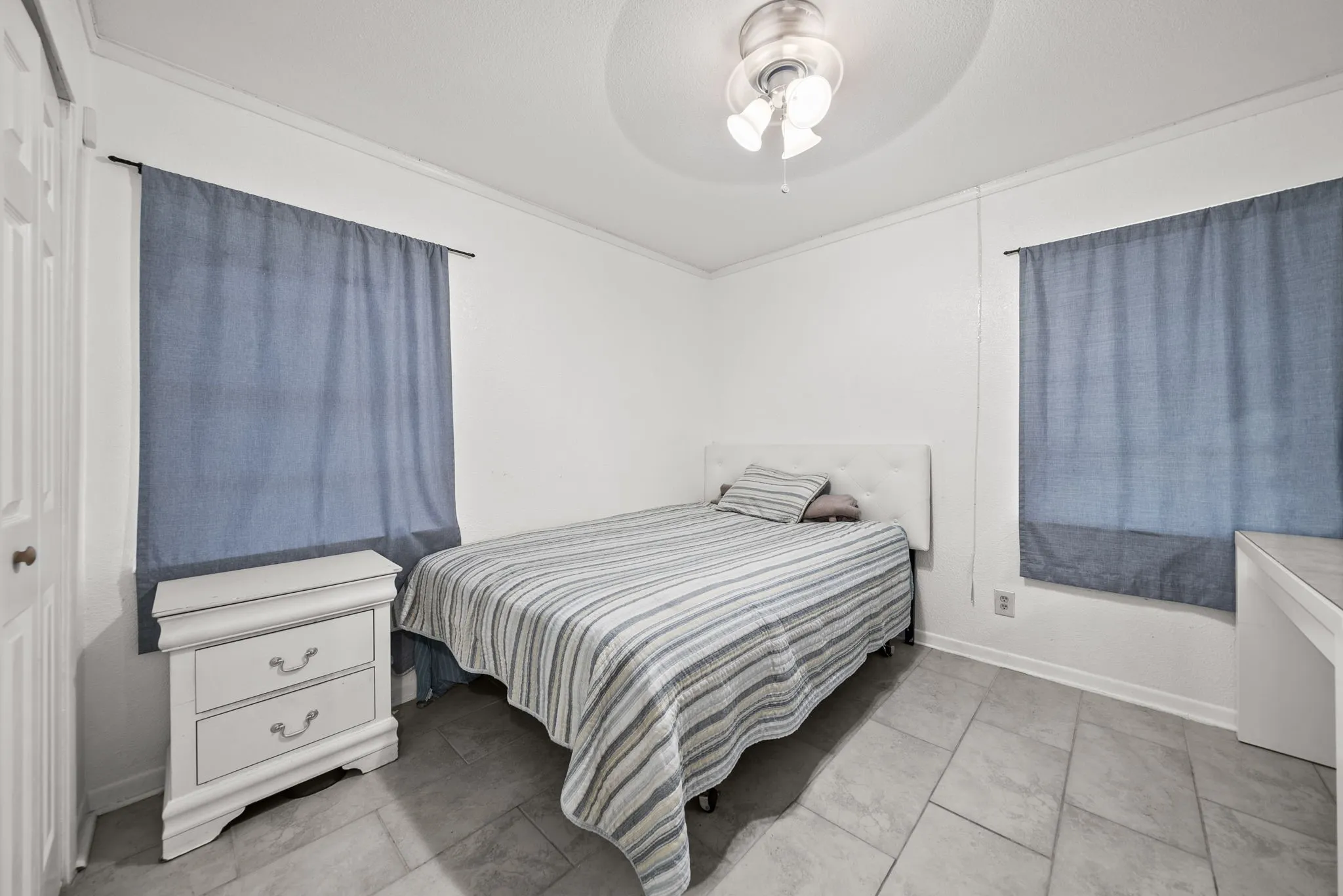 White walls with crown molding, tiled flooring, ceiling fan with light fixture, and two windows with curtain rods