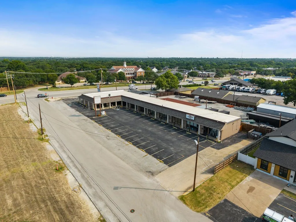 Commercial property featuring a large asphalt parking lot, multiple storefronts with glass entrances, and a brown shingle roof