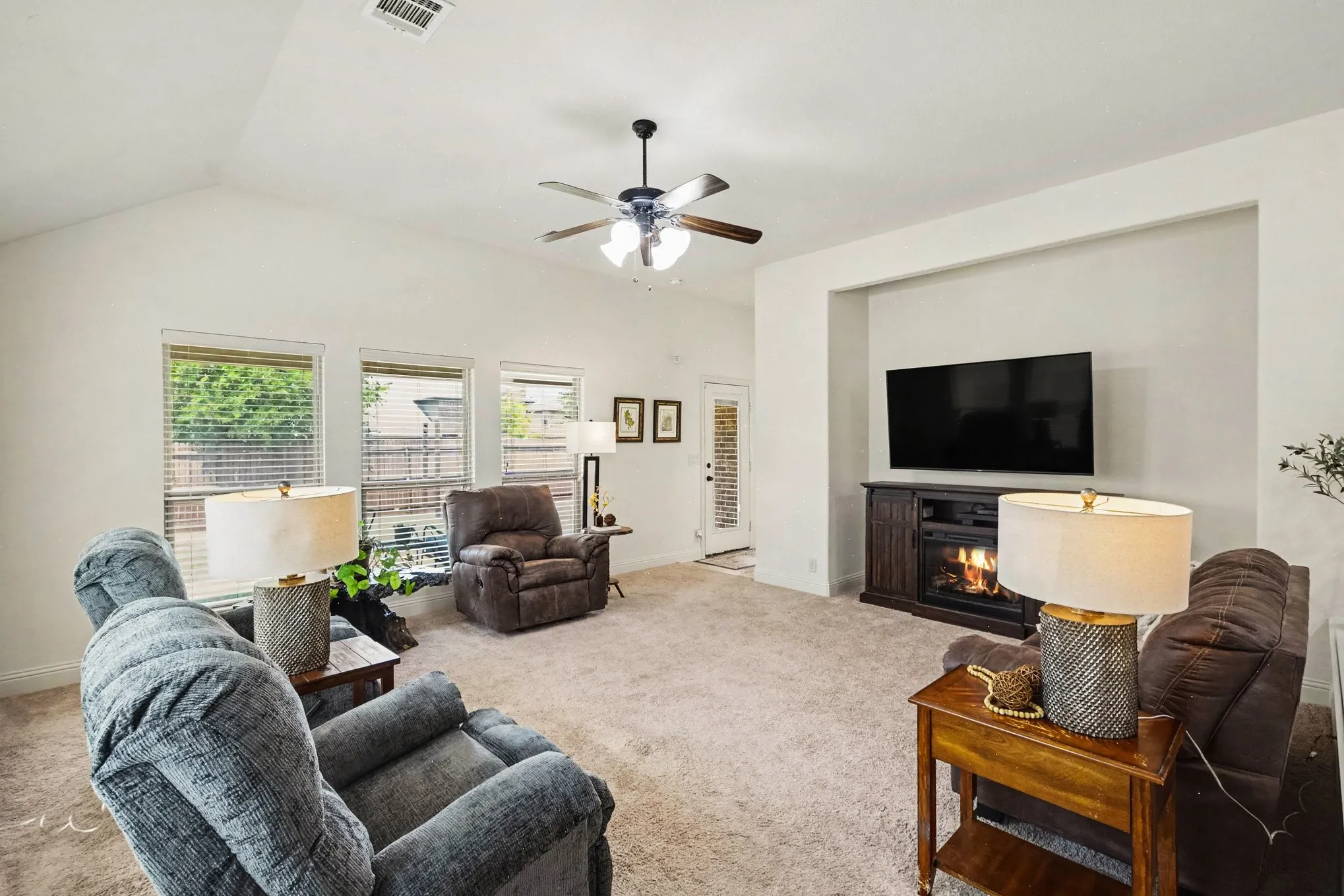 Vaulted ceiling with a ceiling fan, plush carpet flooring, recessed entertainment niche, integrated electric fireplace, and multiple windows with blinds