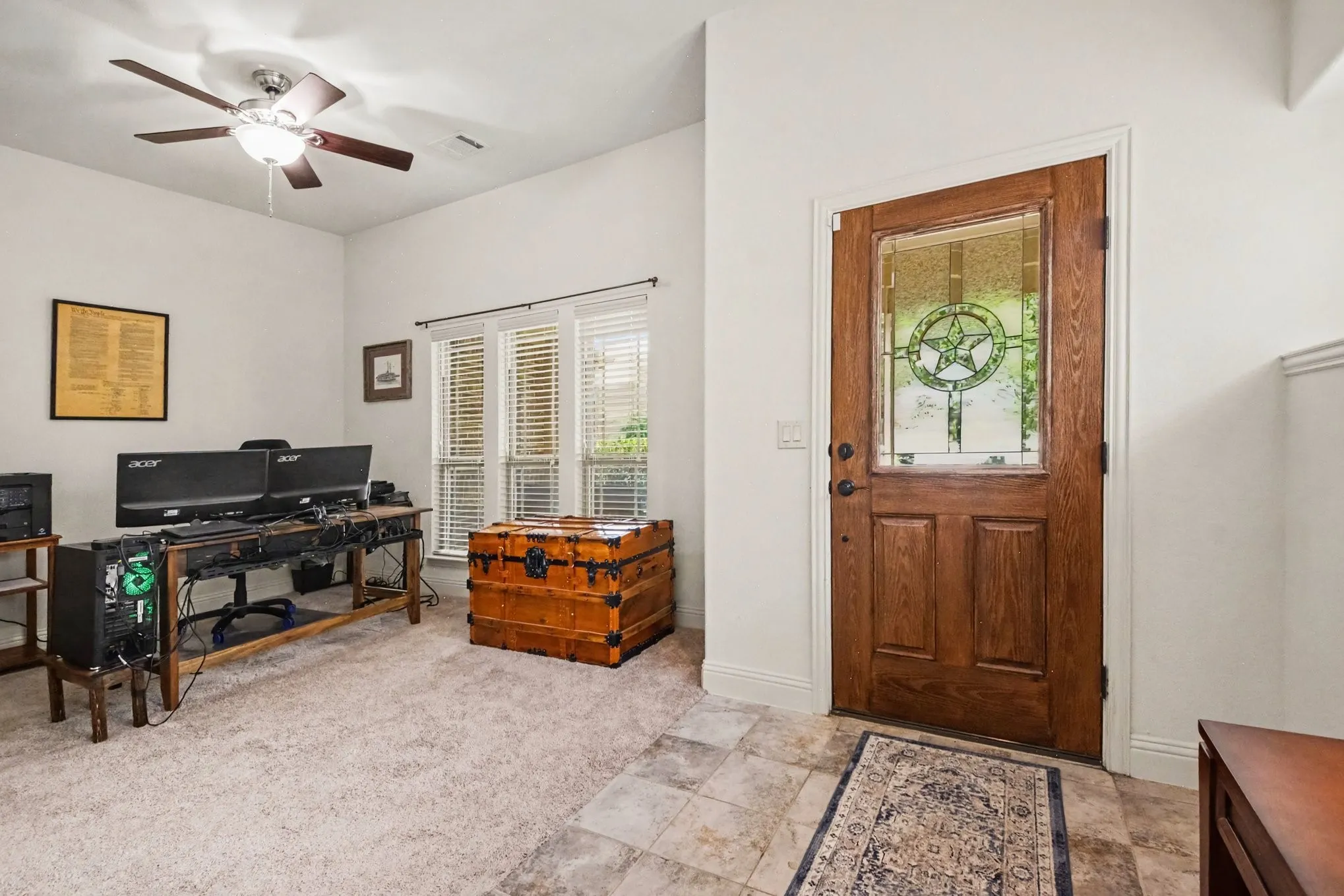 Wood entry door with decorative glass insert, tile flooring, carpeted room, ceiling fan, and multiple windows with blinds