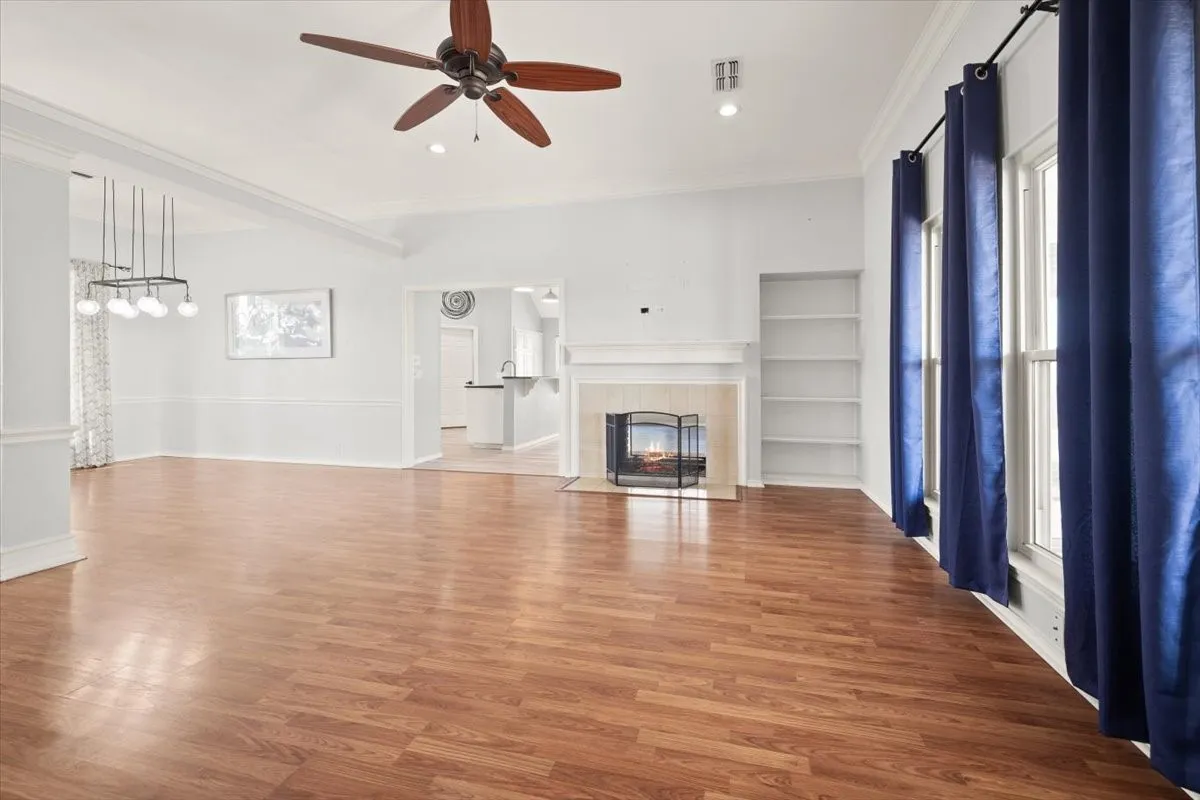 Spacious living area featuring wood-finish flooring, a fireplace with a light-colored mantel, built-in shelving, and a ceiling fan