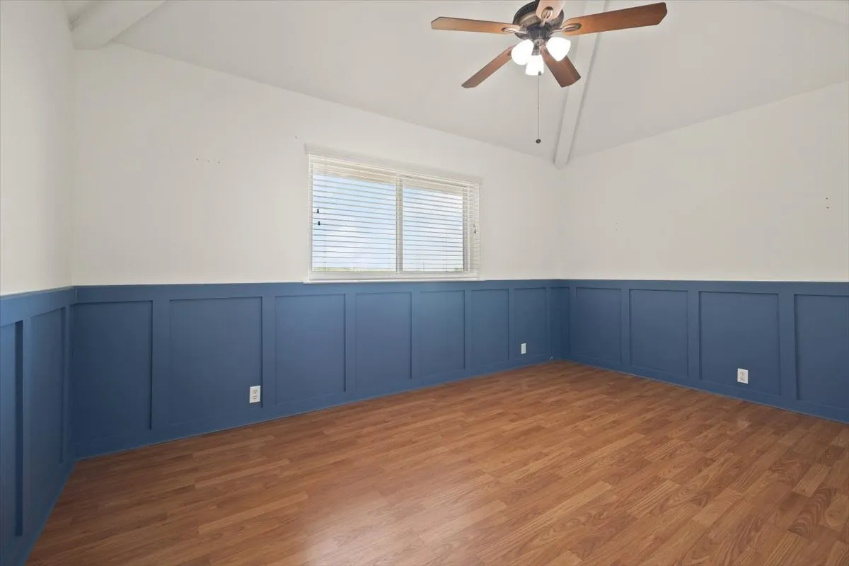 Vaulted ceiling with exposed beam, wood-finish flooring, accent wainscoting, and a ceiling fan with integrated lighting
