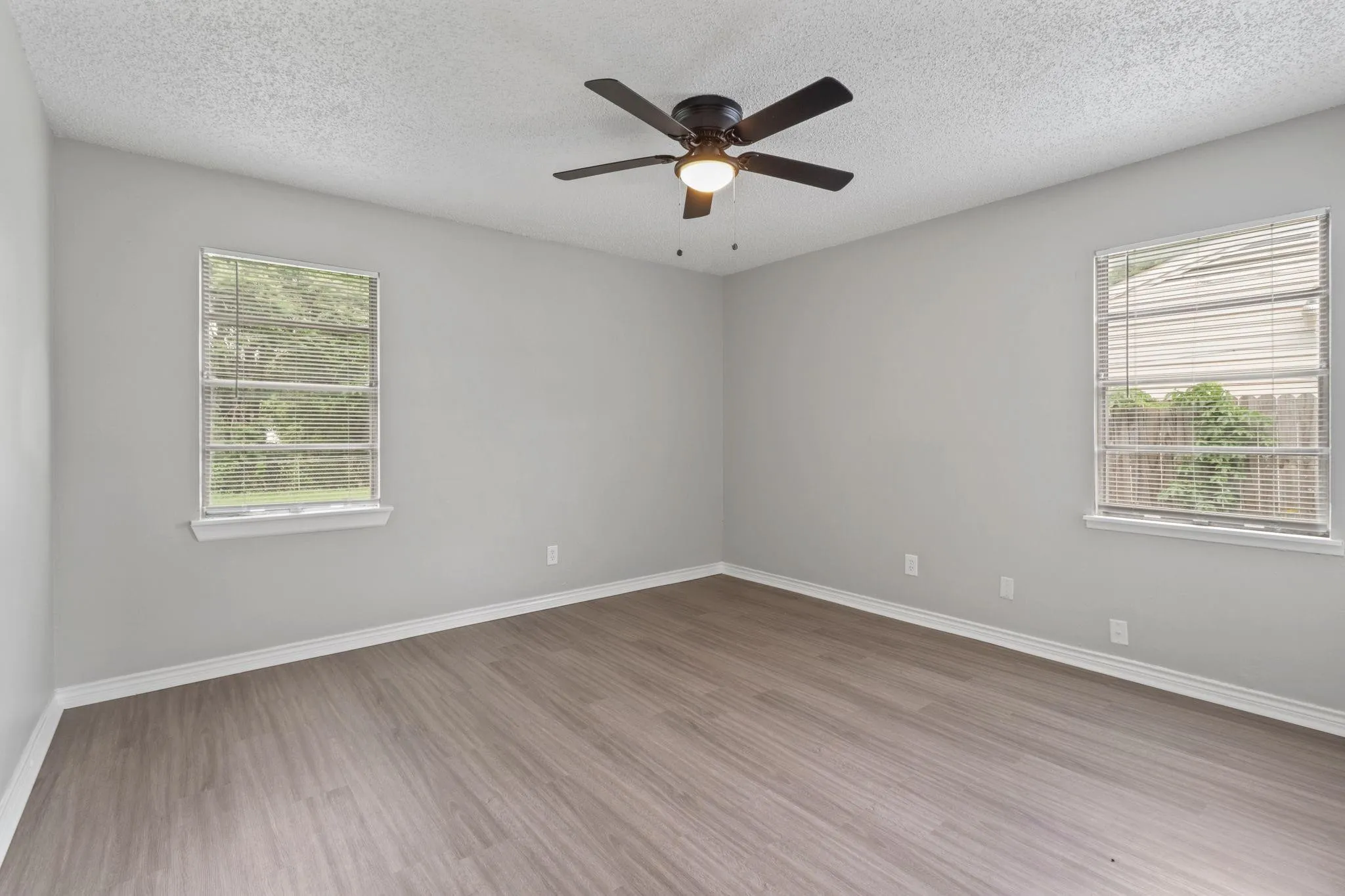 Spacious room featuring wood-finish flooring, two large windows with blinds, a ceiling fan with integrated lighting, and neutral wall paint