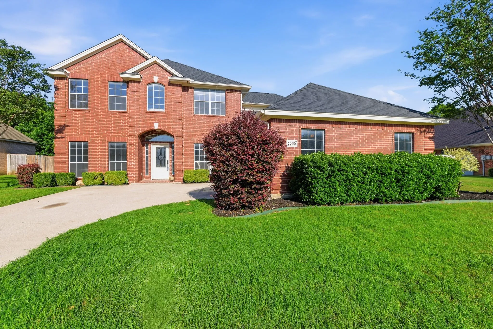 Brick facade with white trim, arched entry, mature landscaping, paved driveway, and gabled roofline