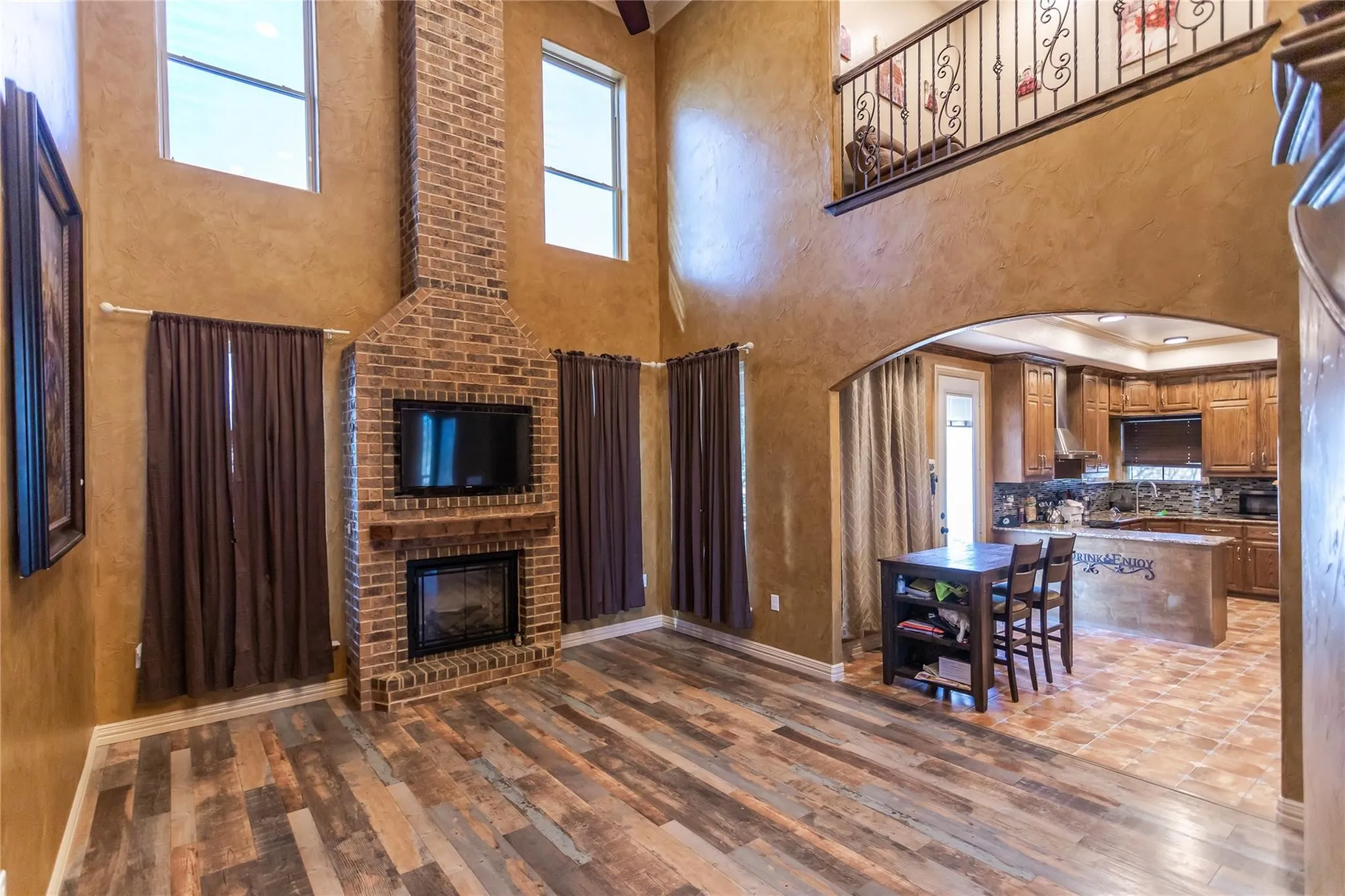 Two-story brick fireplace, wood-finish flooring, arched entryway, open-concept kitchen, and second-story interior balcony with wrought iron railing