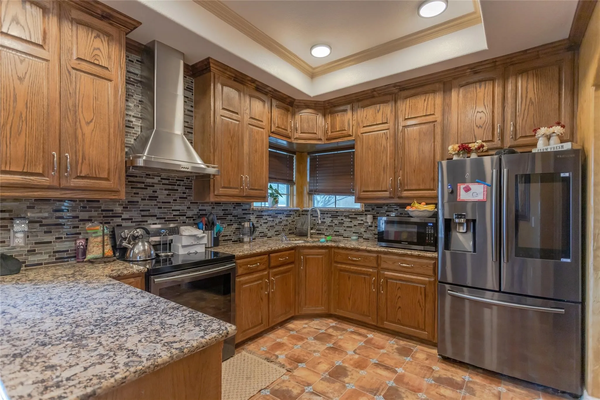 Kitchen featuring wood cabinetry, stainless steel appliances, mosaic tile backsplash, stone countertops, and a tray ceiling with recessed lighting