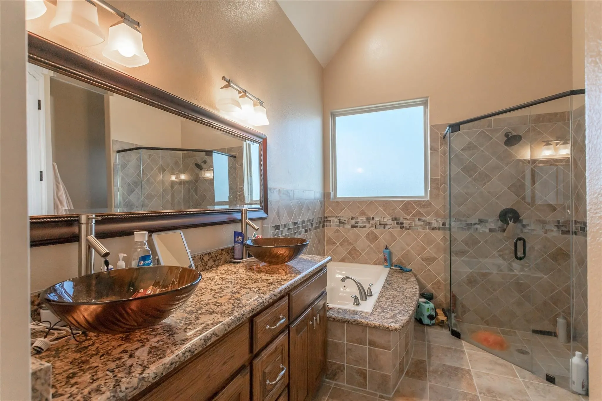 Dual vessel sinks with bronze-finish basins, granite-style countertops, wood-finish cabinetry, and a large soaking tub with tiled surround