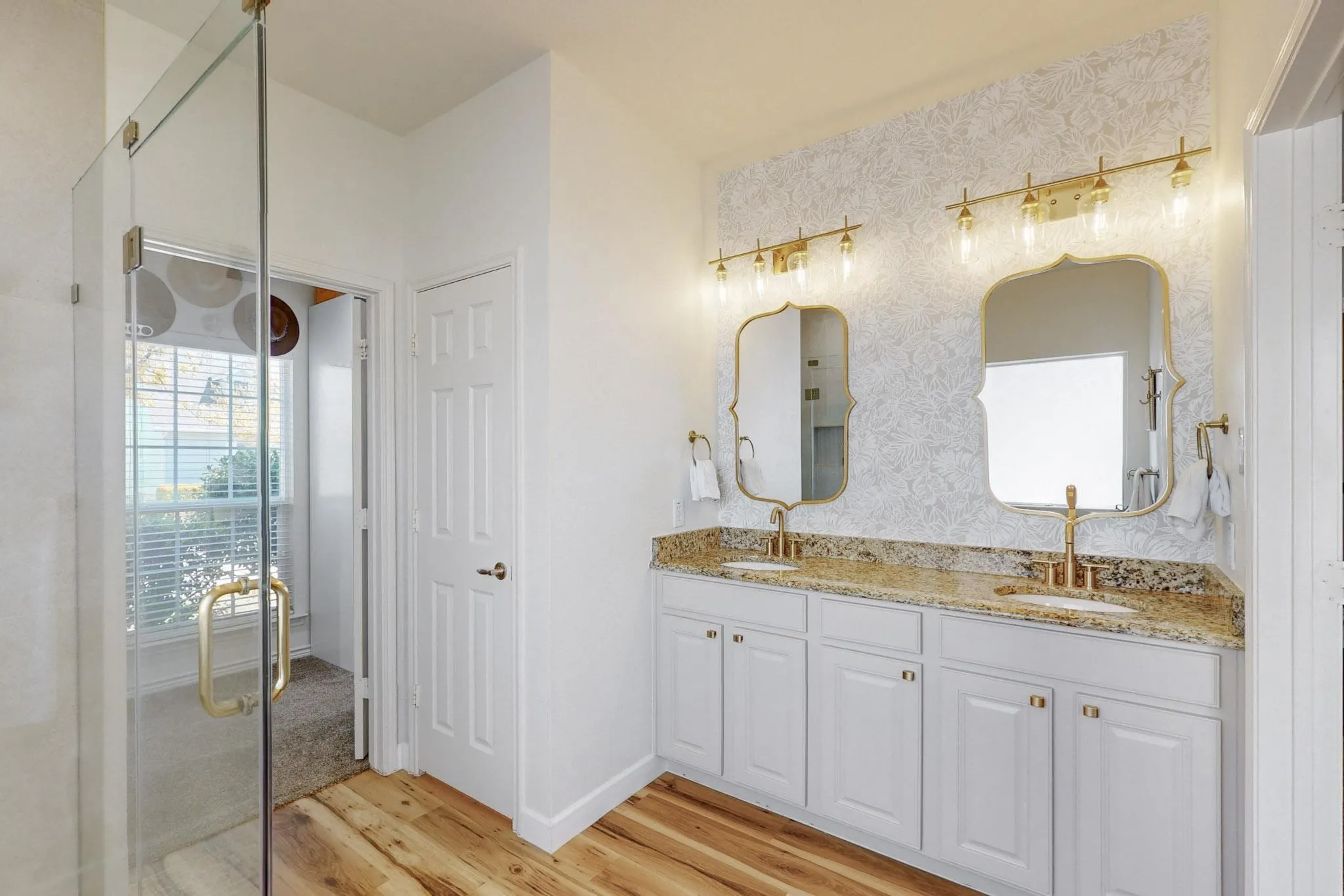 Double vanity with undermount sinks and gold-tone fixtures, framed mirrors with decorative lighting, light wood-finish flooring, and a glass-enclosed shower